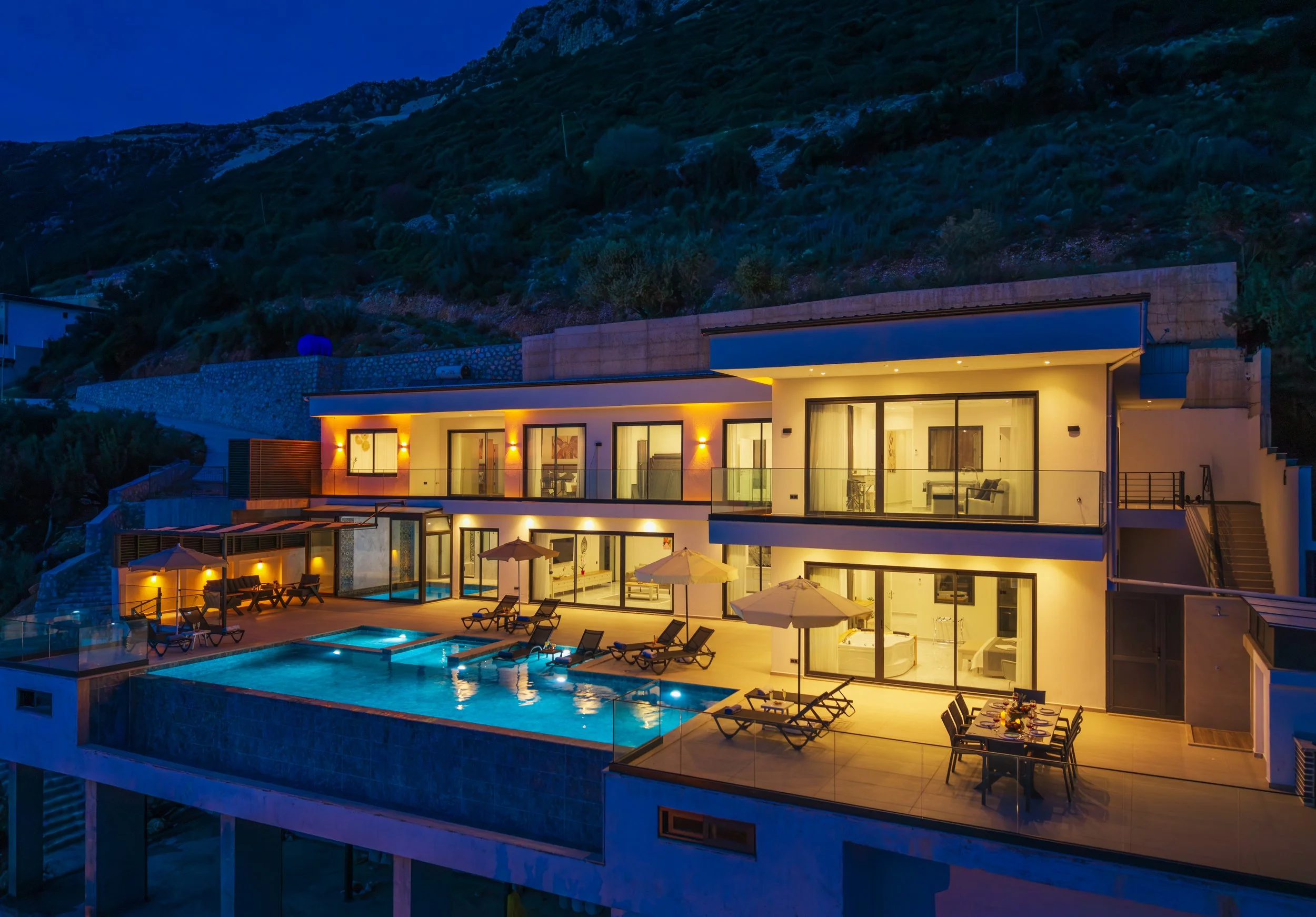 Modern multi-level house at night with lit interior and outdoor pool area, mountain background, and patio furniture