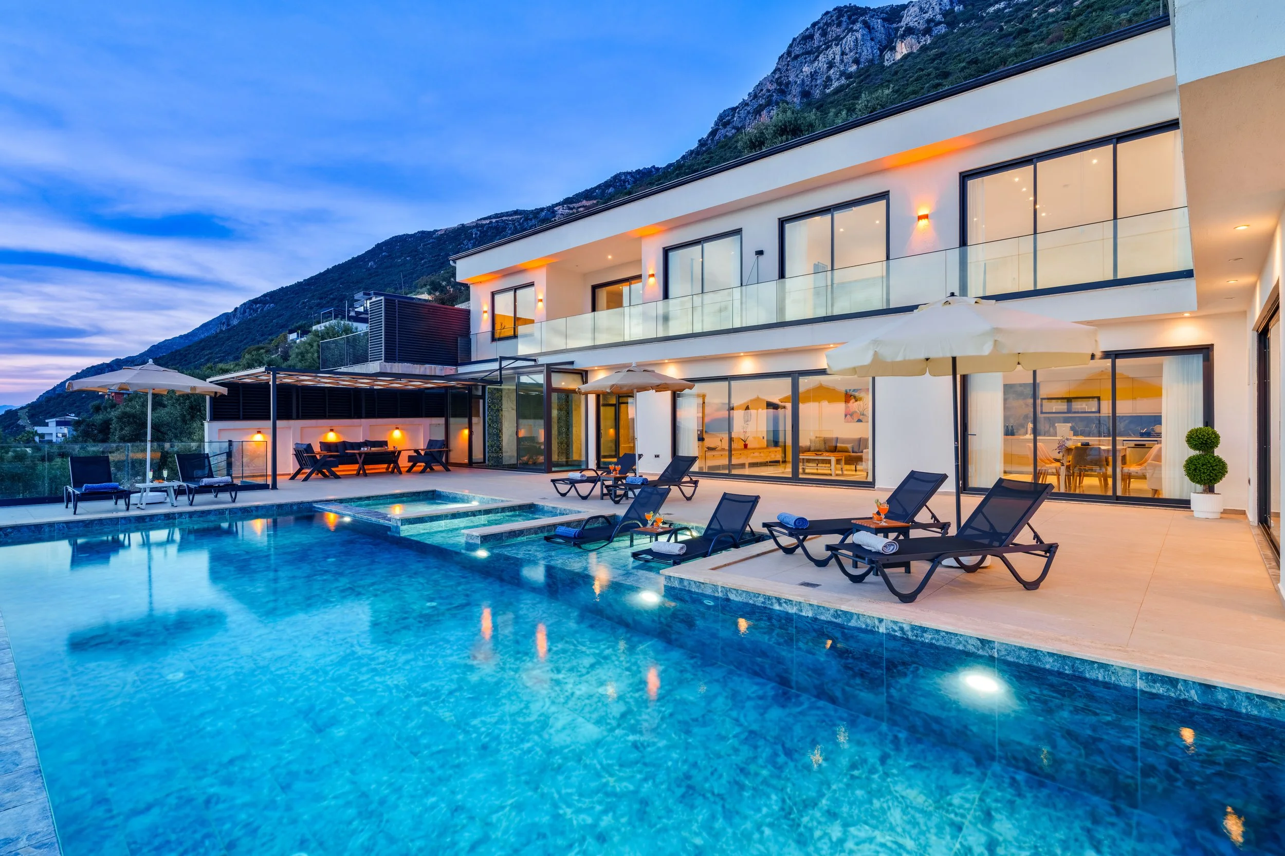 Modern house with large glass windows, outdoor pool area with lounge chairs and umbrellas, mountain backdrop at dusk, warm interior lighting visible through windows.