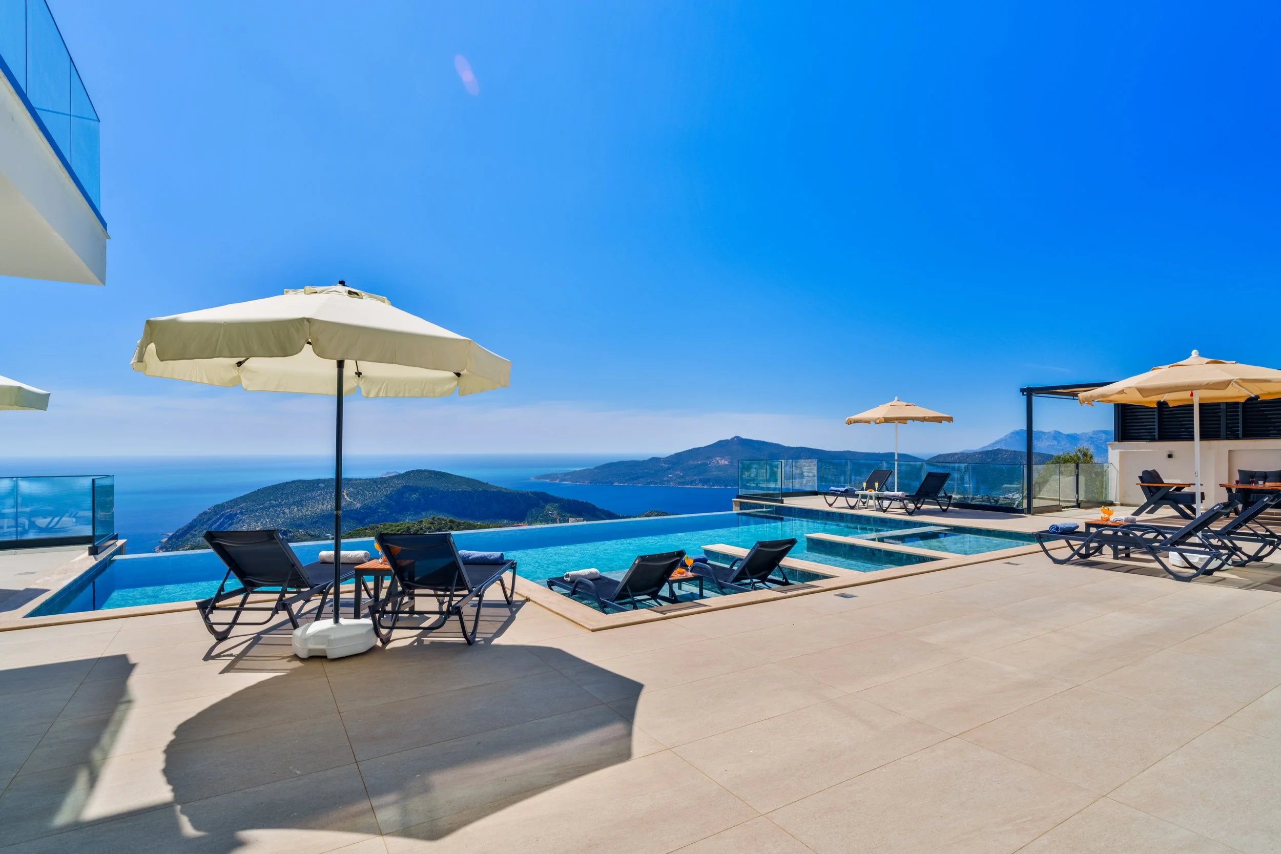 Luxury poolside area with lounge chairs and umbrellas overlooking a scenic mountain and ocean view under a clear blue sky.