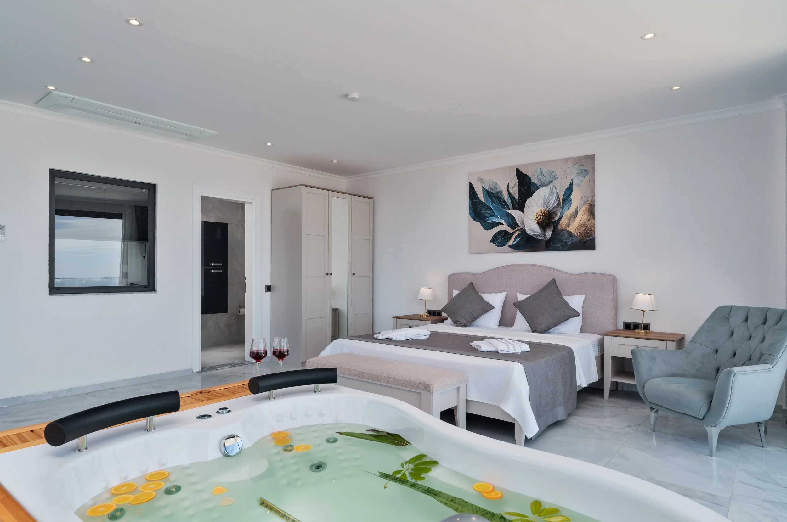 A modern, minimalistic bedroom with a large bed, nightstands, and a wall painting of a flower. There is a window and a door leading to a bathroom, with a jacuzzi tub filled with water and floating leaves and orange slices in the foreground.