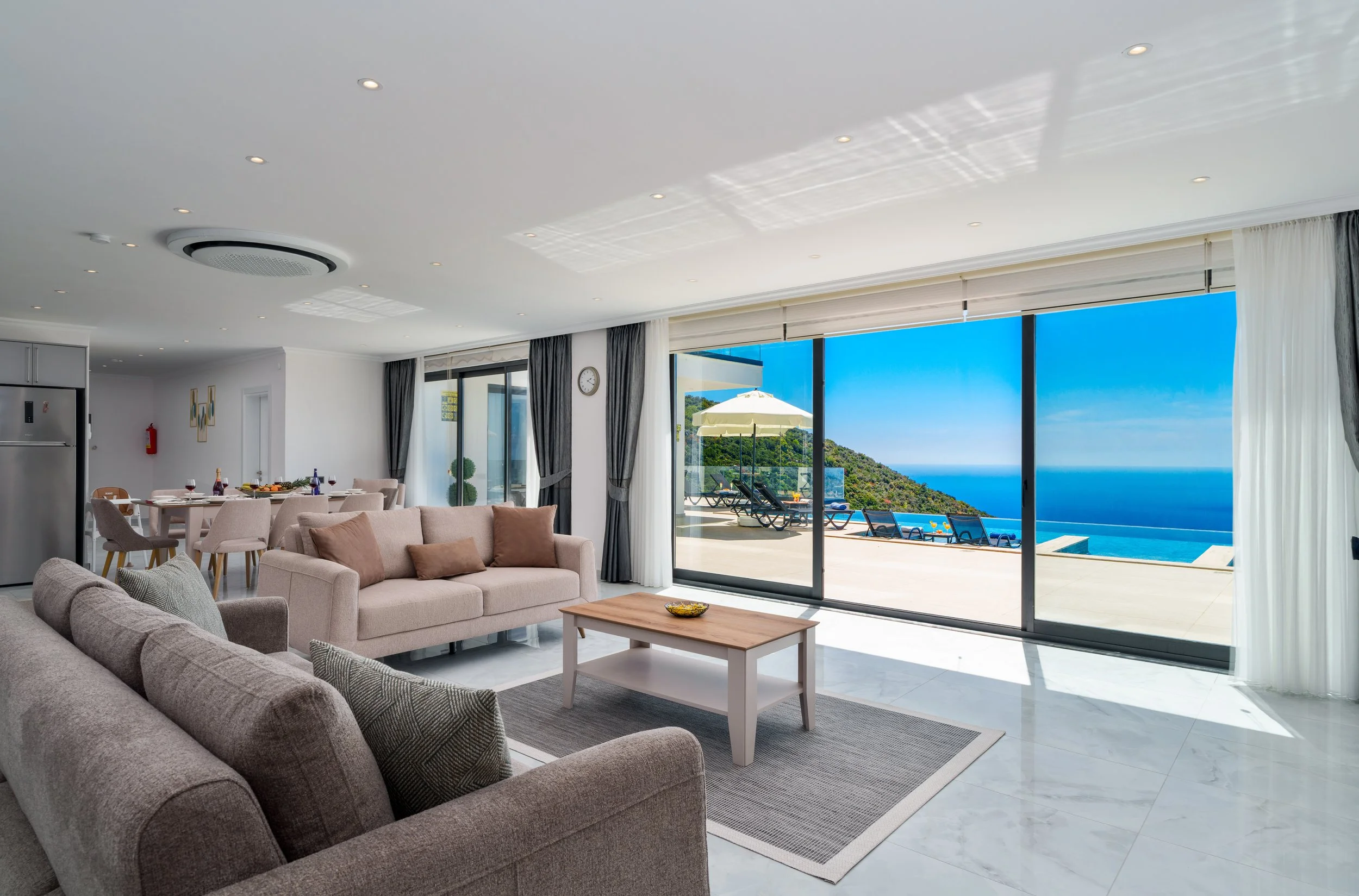 Living room with large glass sliding doors showing a view of the ocean and greenery outside, with patio furniture and a swimming pool.