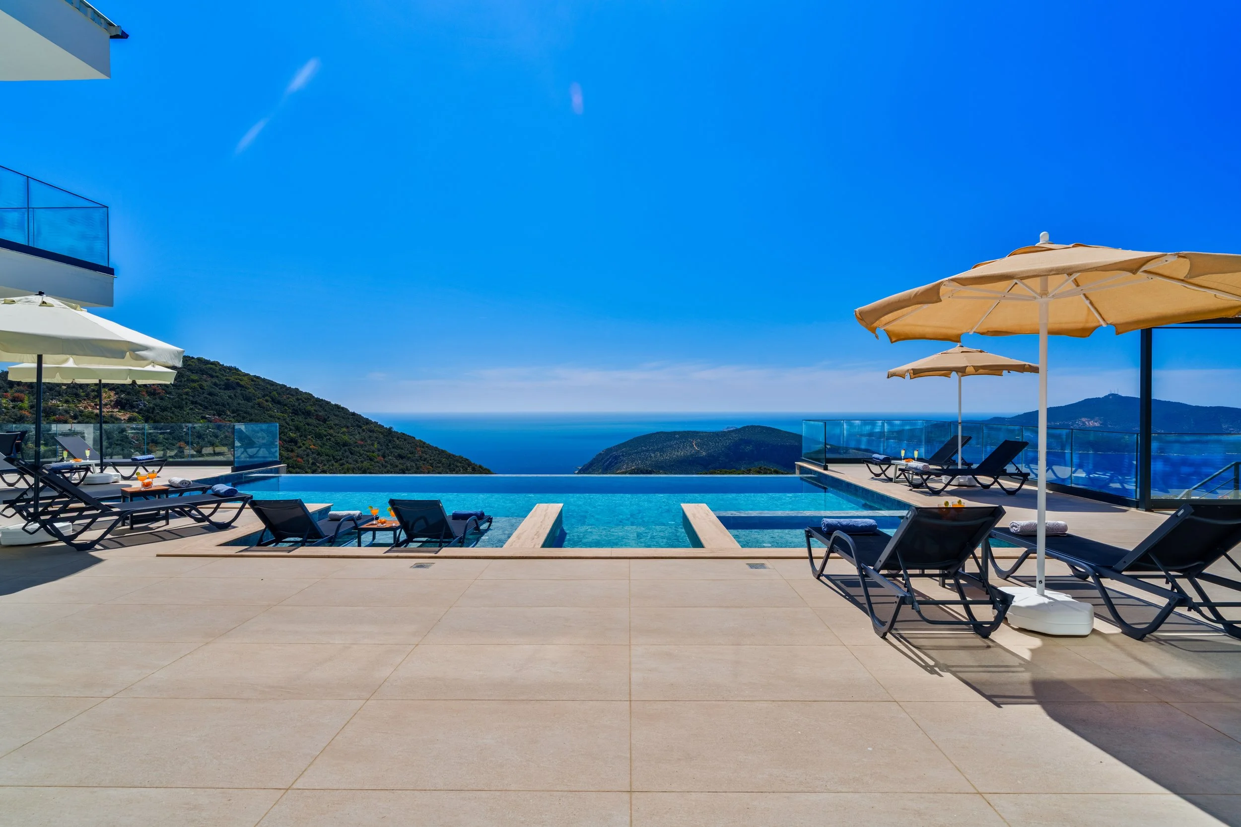 View of a modern infinity pool with lounge chairs and umbrellas on a terrace overlooking a mountainous landscape and the ocean on a clear, sunny day.