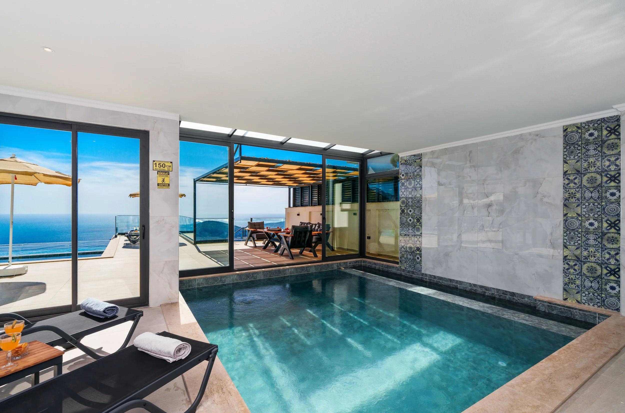Indoor swimming pool area with lounge chairs, towels, and a view of the ocean through glass walls and sliding doors.