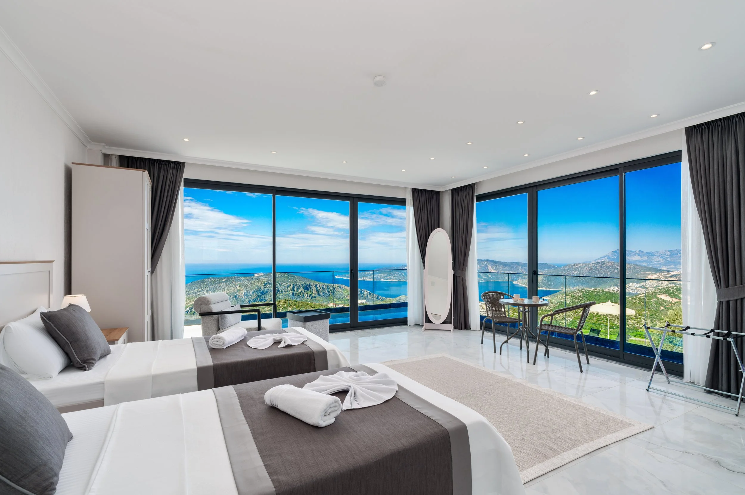 A modern hotel room with two beds, sea view through large floor-to-ceiling windows, and a small seating area with a table and chairs.