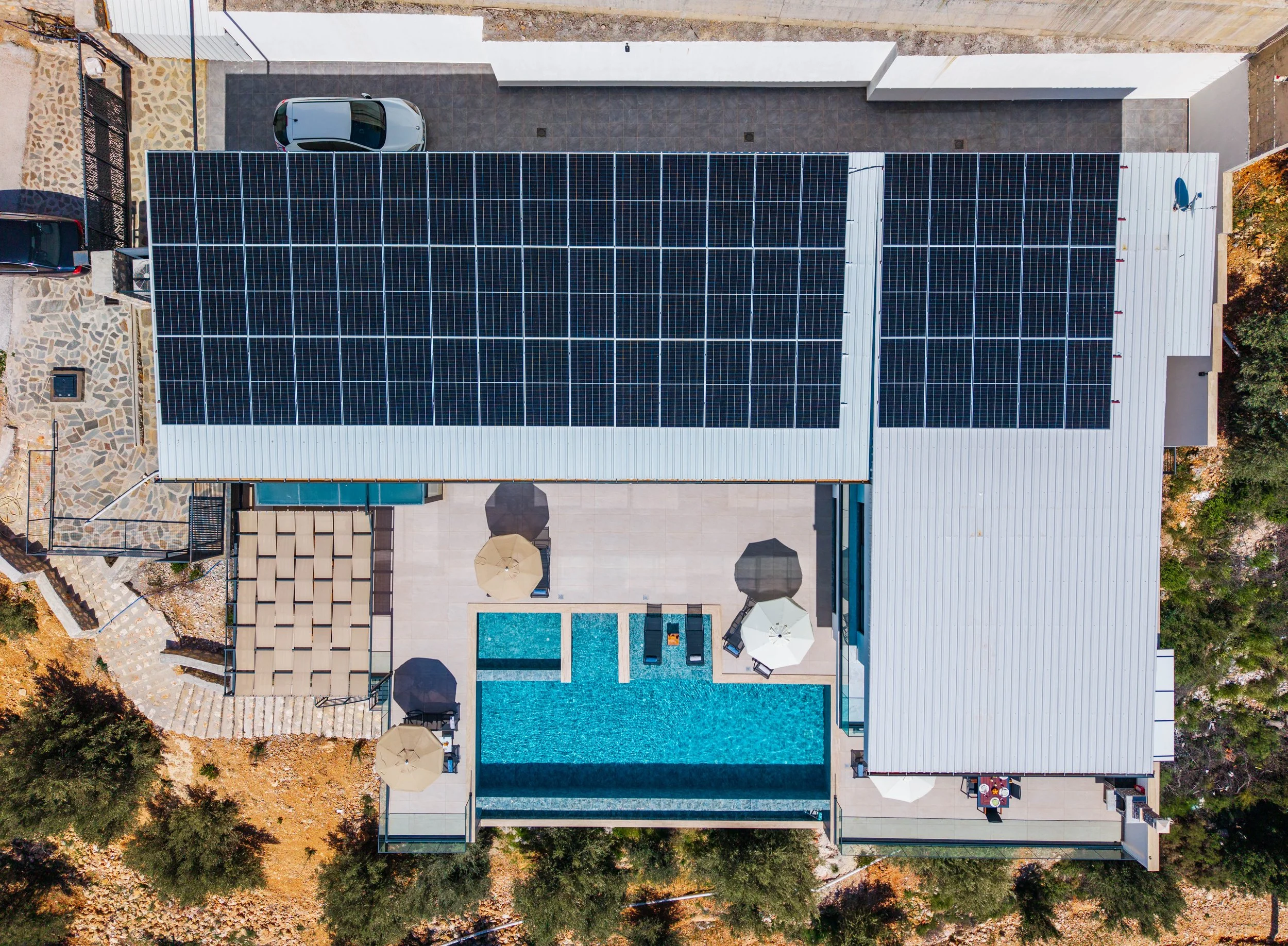 Aerial view of a residential property with solar panels on the roof, a swimming pool, patio area with umbrellas, and two parked cars, surrounded by trees and landscaped yard.