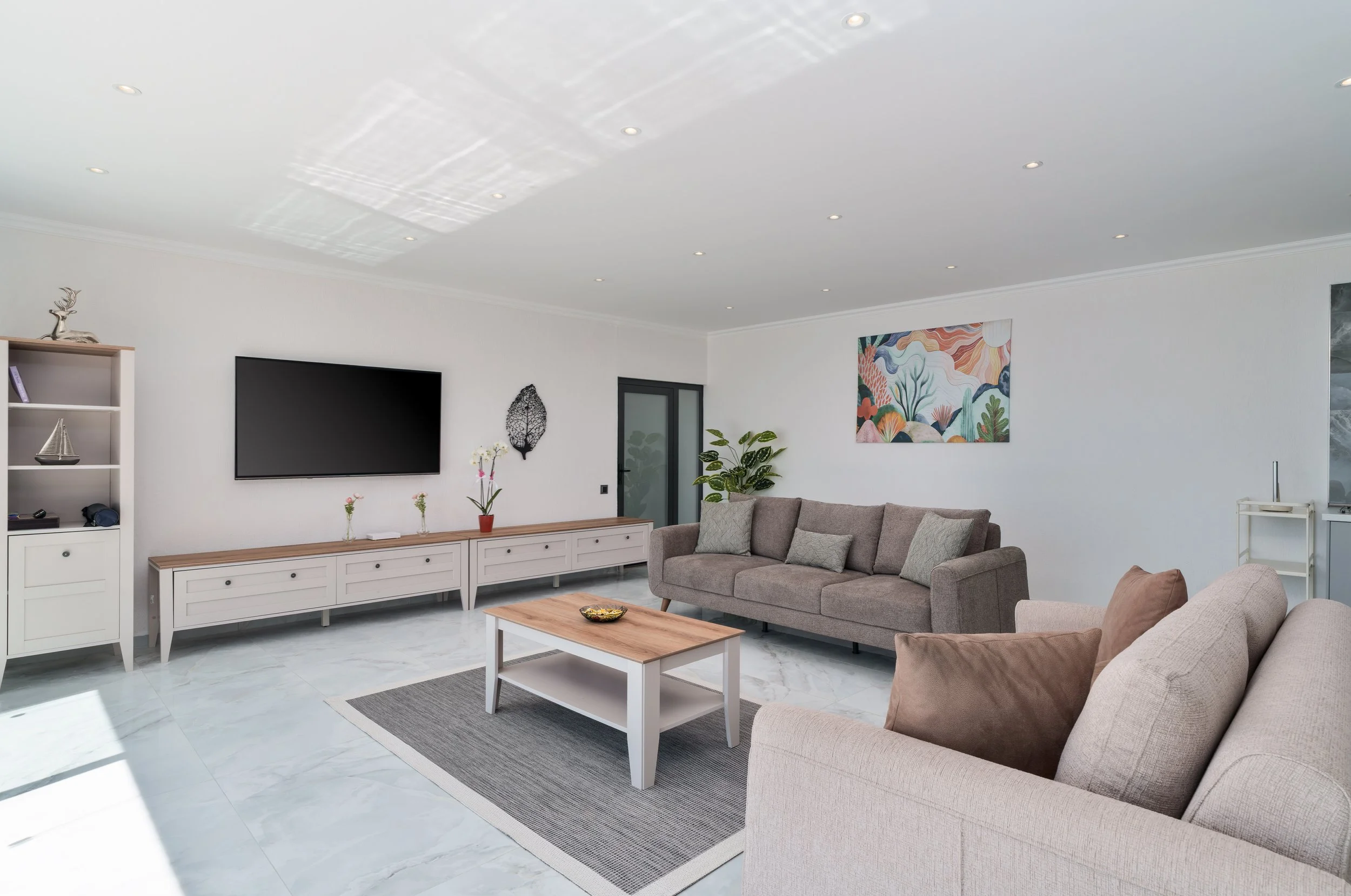 Living room with white walls, a mounted flat-screen TV, a white sideboard with decorative items, a gray sofa with cushions, a wooden coffee table, a colorful abstract painting, and potted plants, illuminated by ceiling lights.