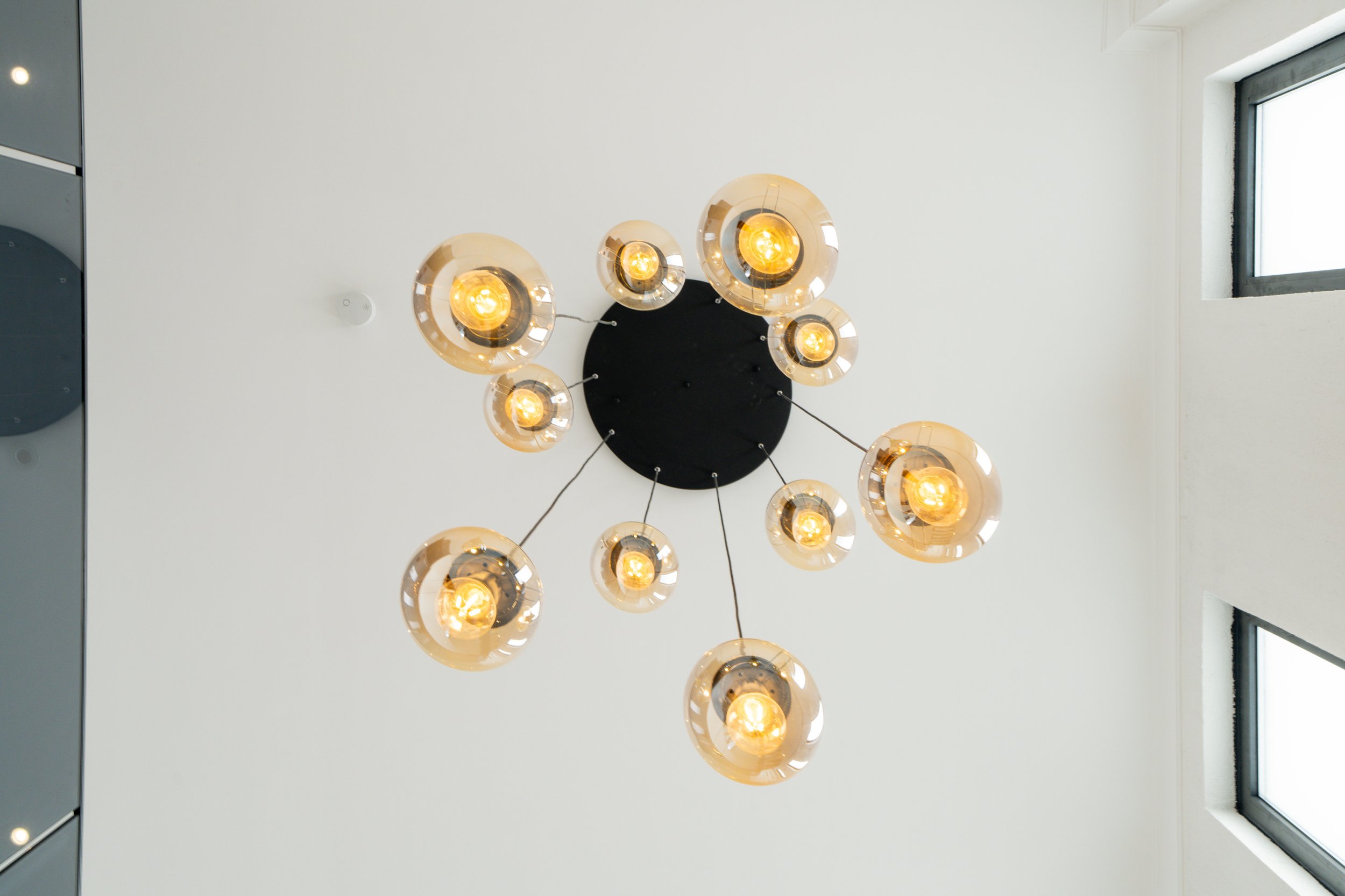 Modern ceiling light fixture with multiple glass globes emitting warm light, attached to a black circular base on a white ceiling near a window.