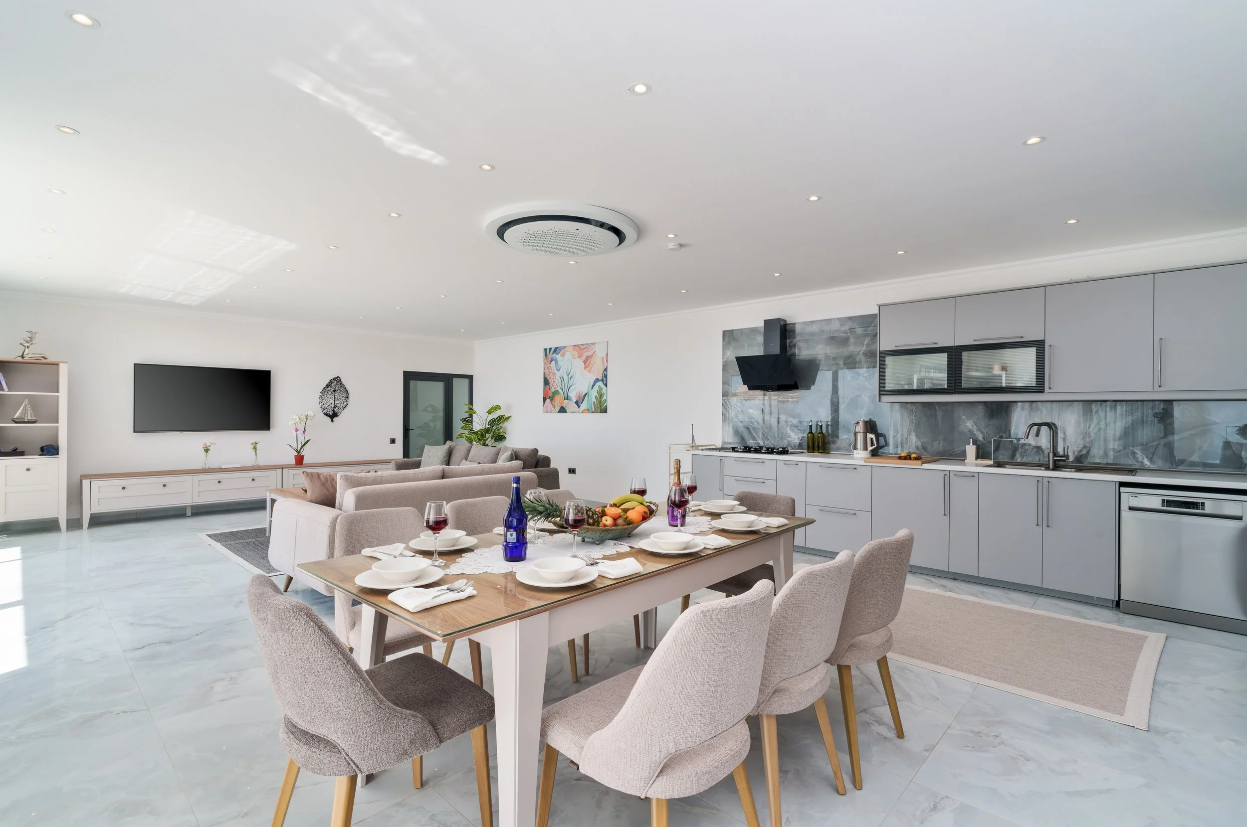 Open-concept living room and kitchen with a dining table set for dinner. The room features white walls, marble floors, a wall-mounted TV, and modern kitchen cabinets with a gray backsplash. Decorations include a colorful painting, potted plants, and 