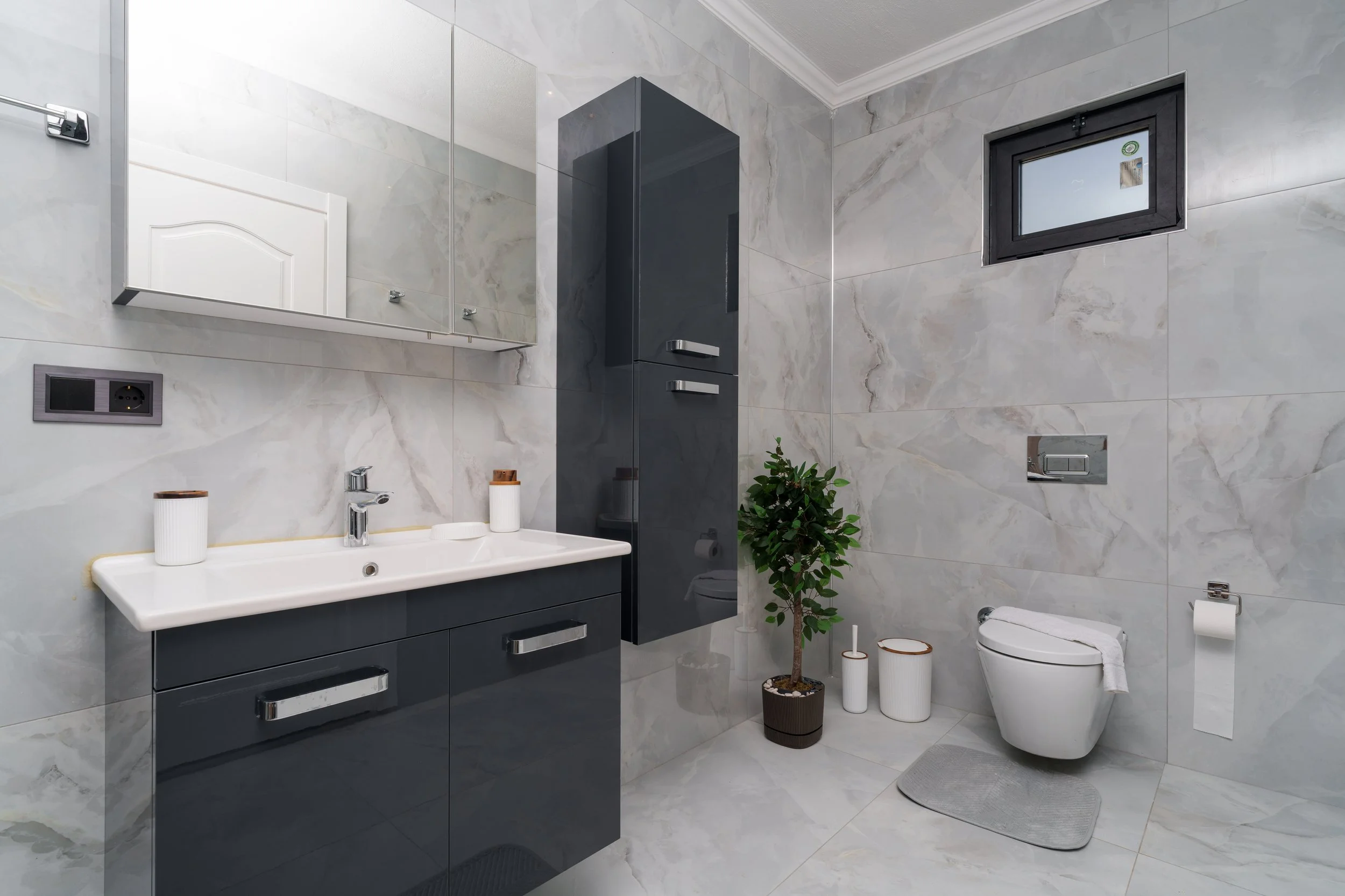 Modern bathroom with marble walls, a sink vanity with dark cabinets, a mirror, a small window, a toilet, a bidet, potted plant, and white containers.