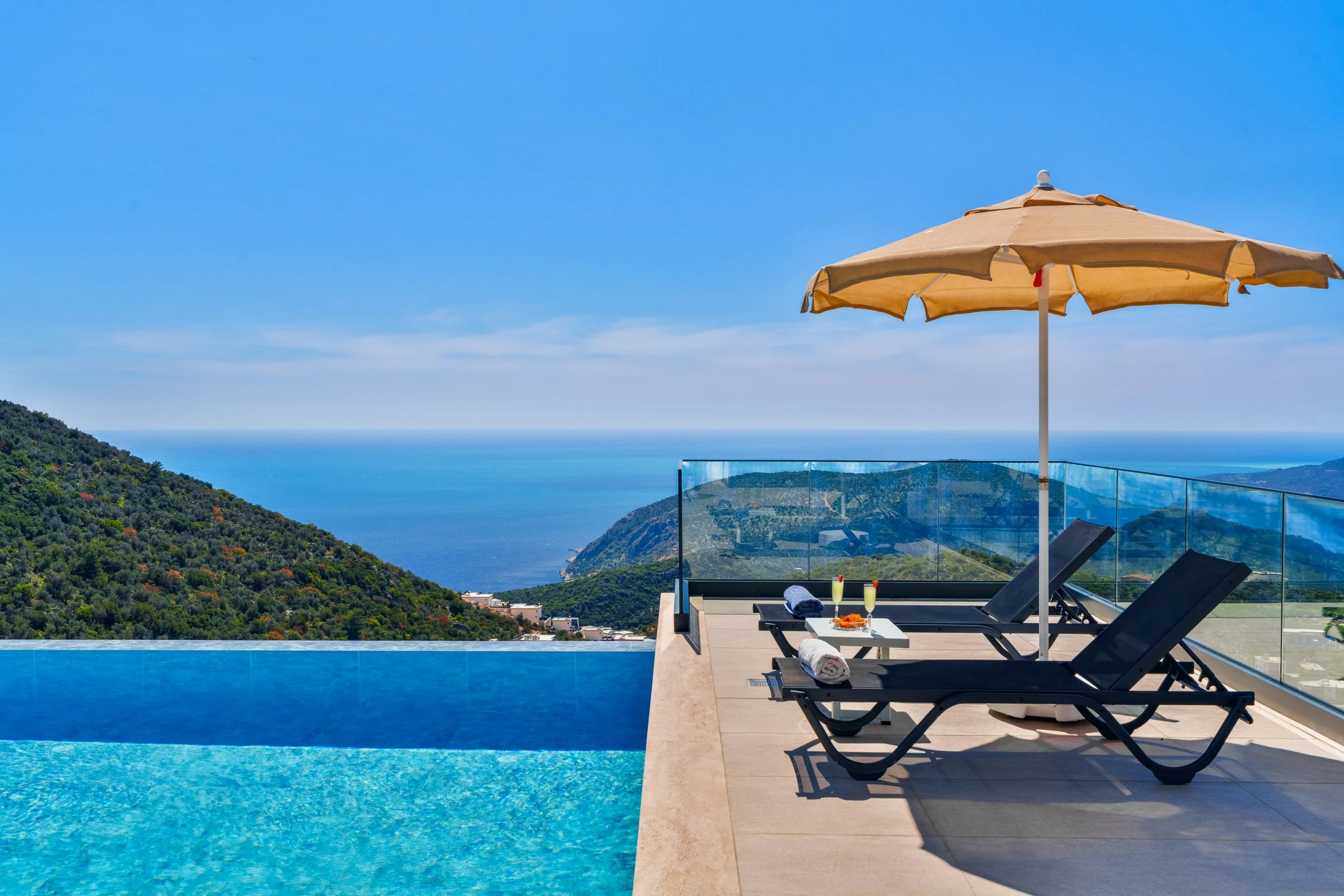 Luxury outdoor pool with view of hillside and ocean, lounge chairs with towels, a large beige umbrella, drinks, and glasses on a table.