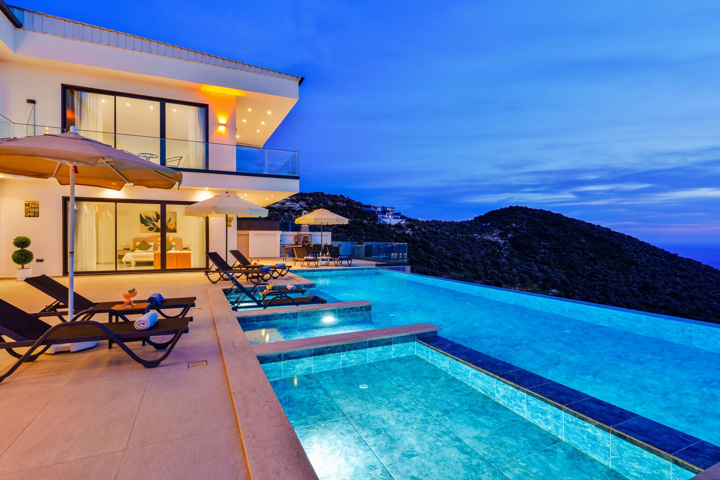Luxury modern house overlooking a mountain landscape at dusk with outdoor pool, lounge chairs, umbrellas, and warm lighting.