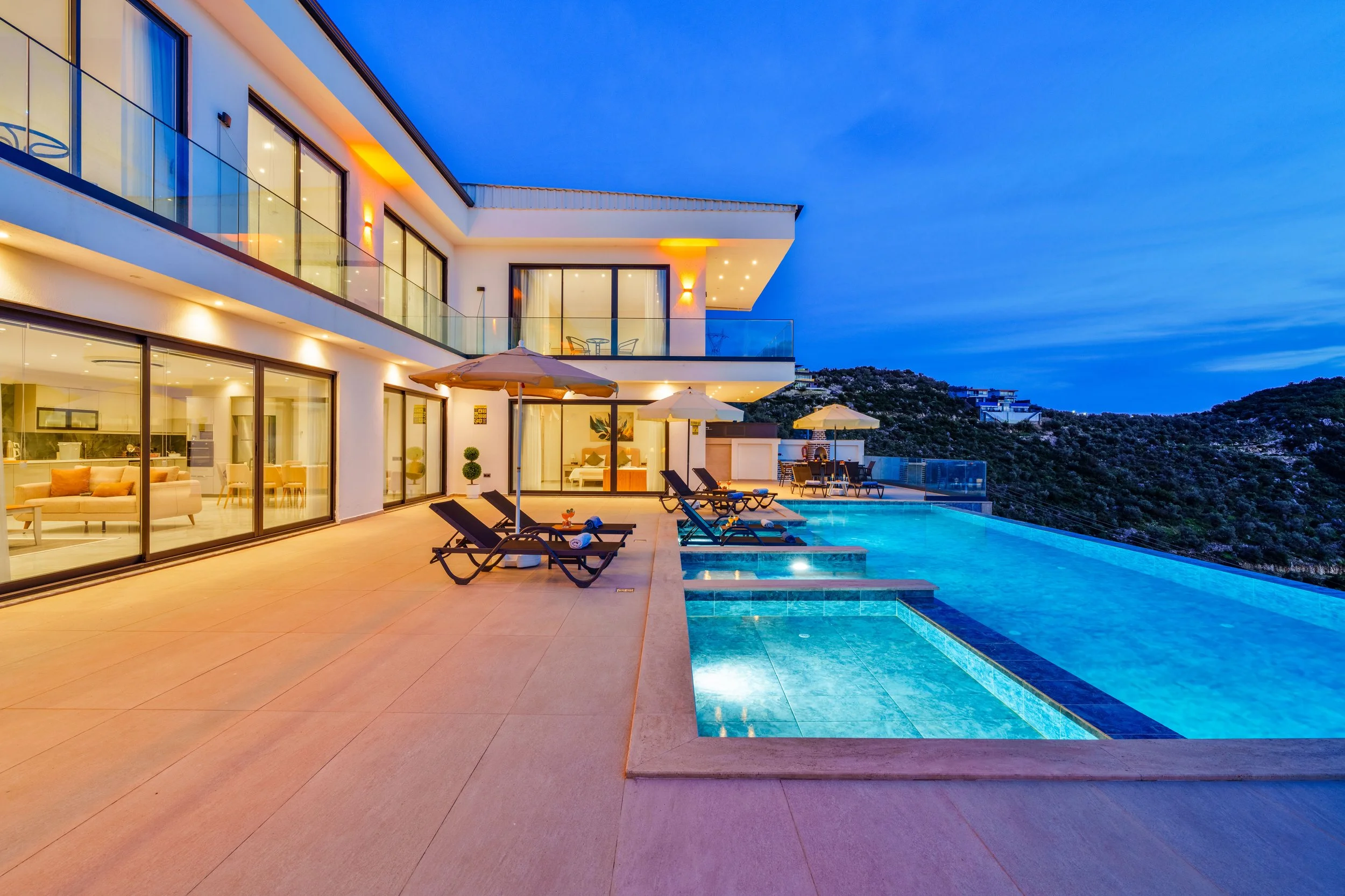 A modern multi-story house with large glass windows and balconies, featuring a swimming pool with lounge chairs and umbrellas on a patio, set against a hillside during the evening with a clear blue sky.