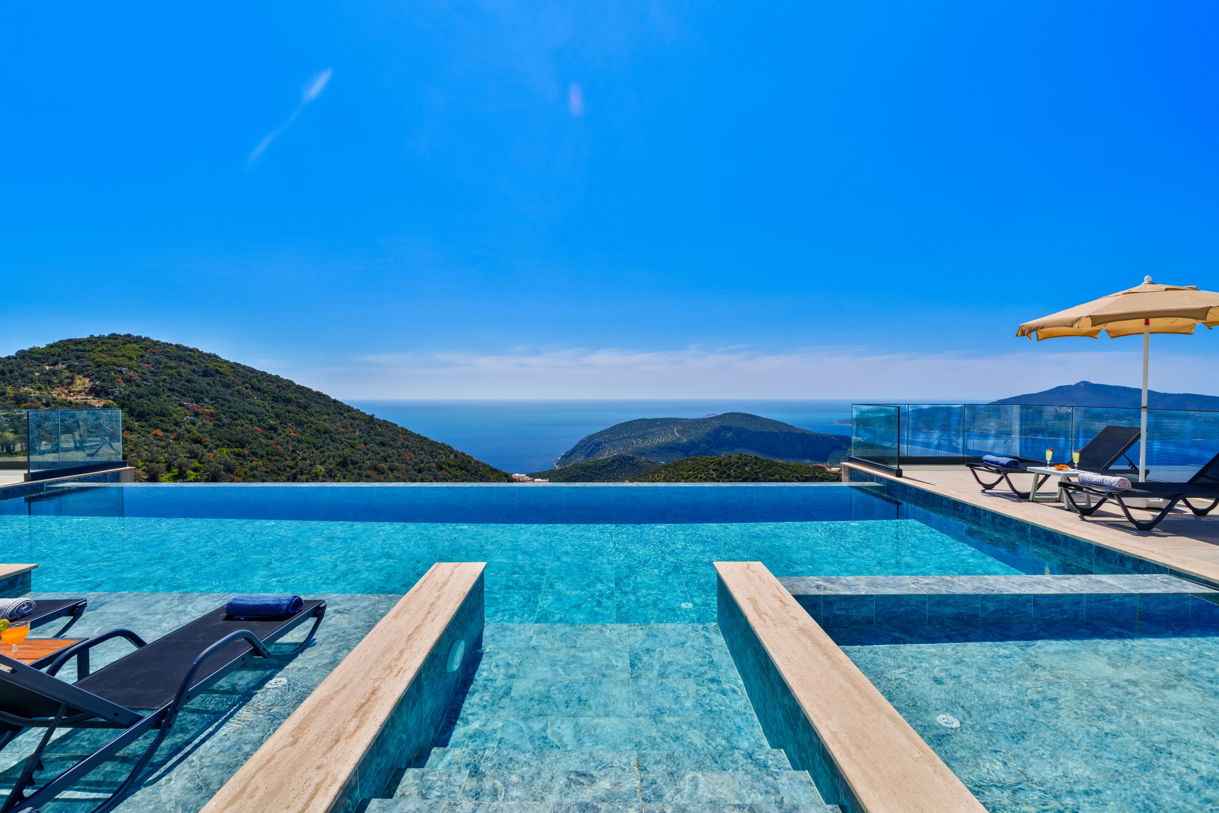 A swimming pool with a view of green mountains and a distant ocean, with lounge chairs and an umbrella beside the pool.