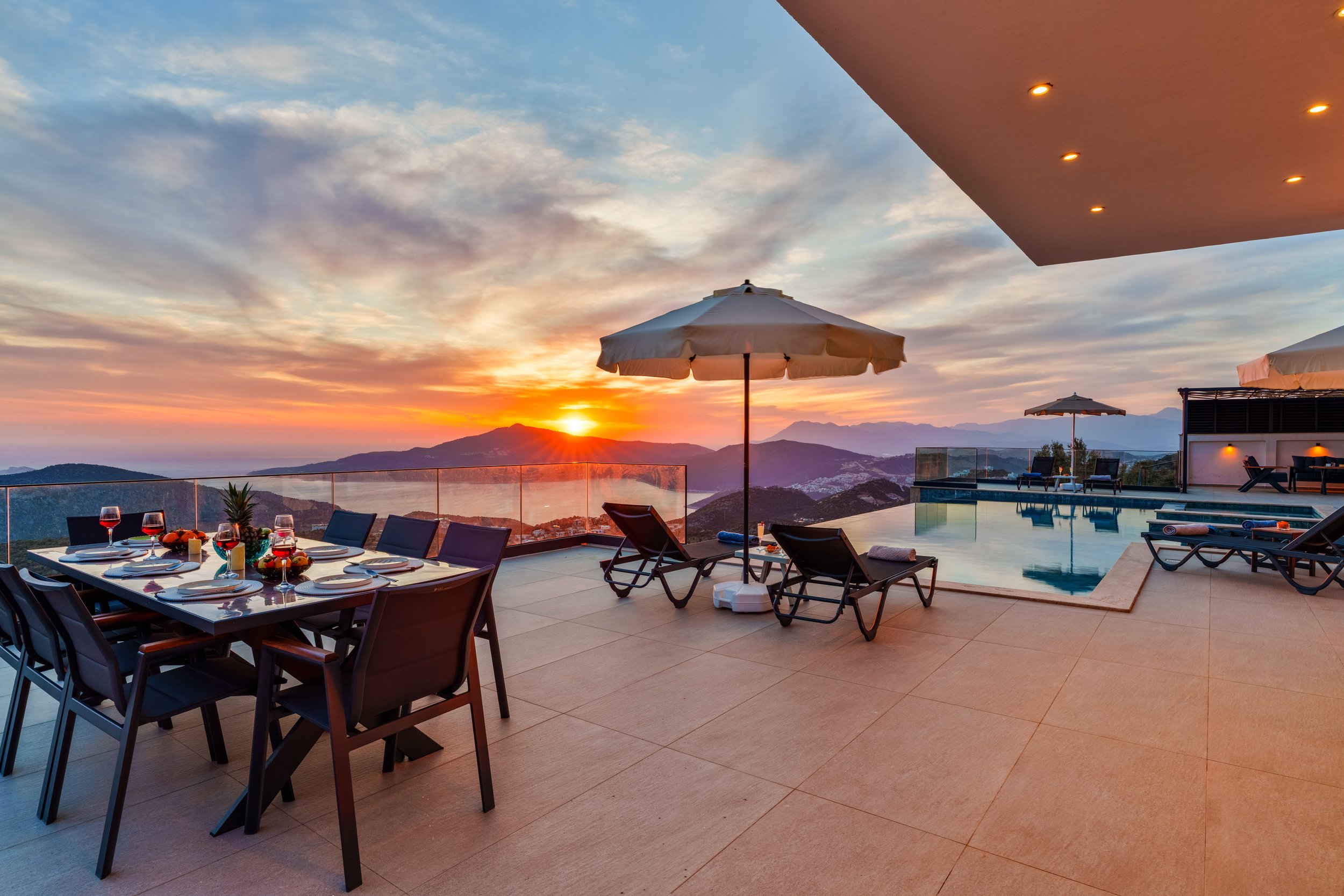 A luxurious outdoor patio at sunset with a dining table set for a meal, lounge chairs, umbrellas, and a swimming pool, overlooking mountains and a colorful sky.