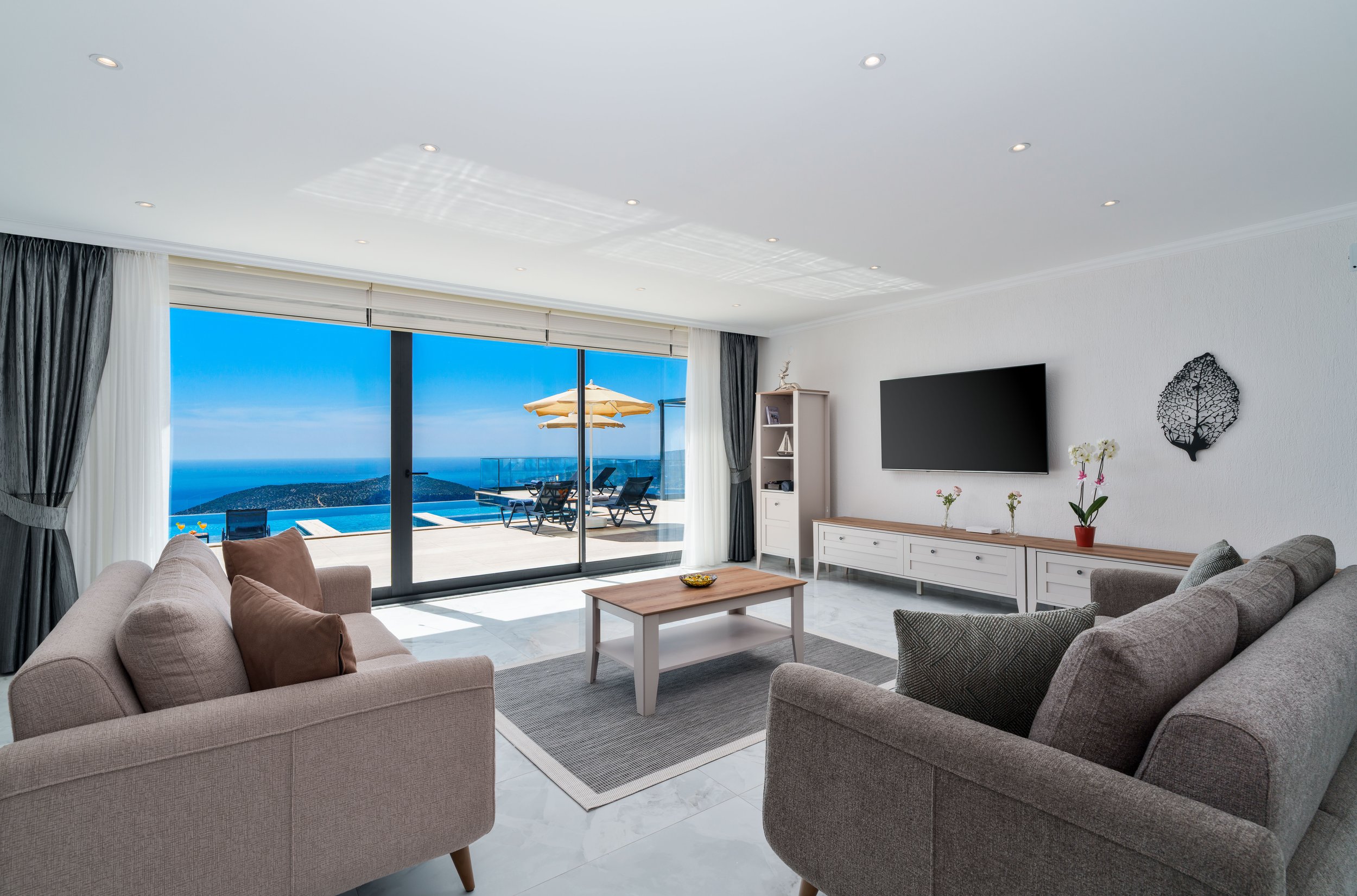 Living room with beige and gray sofas, a white coffee table, a wall-mounted TV, and a large glass sliding door leading to a balcony with an ocean view and outdoor patio furniture and umbrella.
