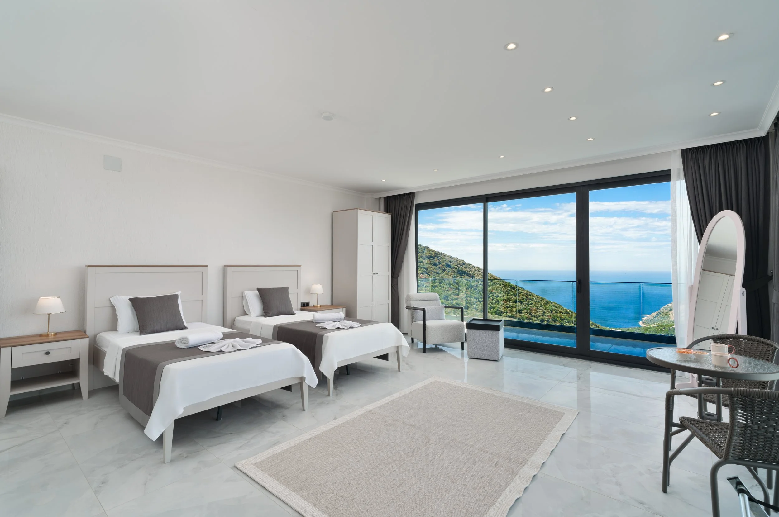 A modern hotel room with two single beds, white headboards, dark gray bedding, and pillows. The room has large sliding glass doors that open to a balcony with a view of the ocean and green hills. There are two nightstands with lamps, a white wardrobe