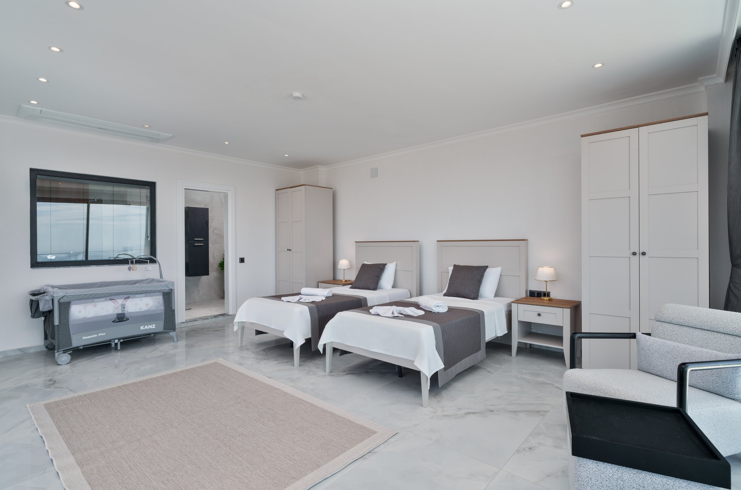 A modern, minimalistic hotel room with three beds, bedside lamps, a wardrobe, a large window, a baby crib, and a sitting area with a gray armchair. The room has white walls, a light-colored marble floor, and ceiling lights.