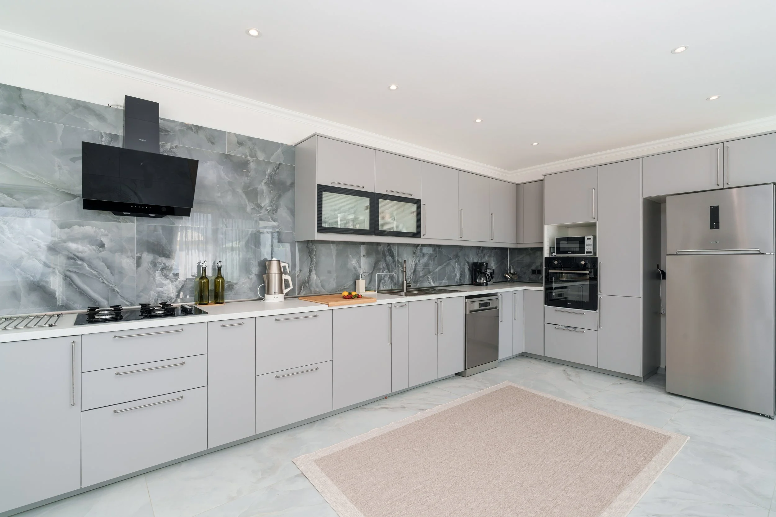 Modern kitchen with gray cabinets, marble backsplash, built-in microwave and oven, stainless steel refrigerator, and a beige rug on white marble floor.