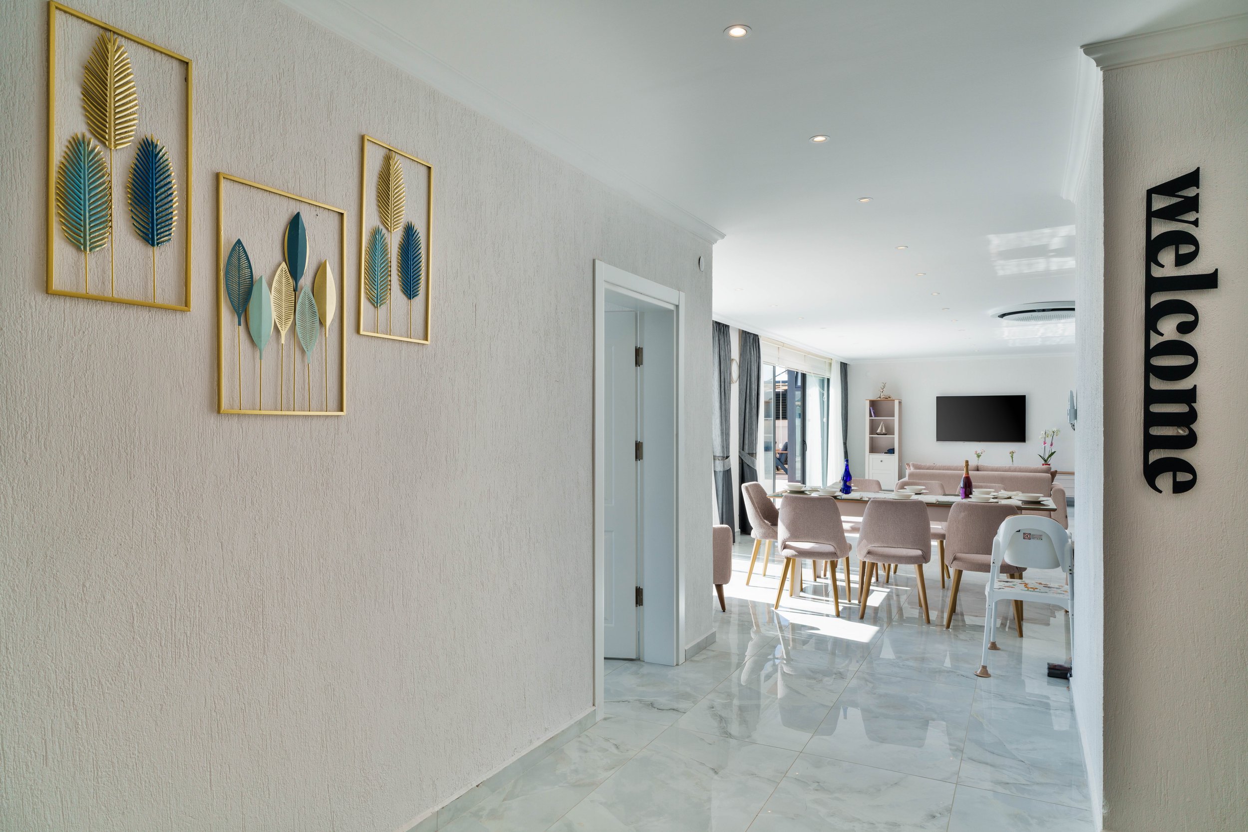 Modern dining area with pastel pink chairs arranged around a white dining table set with plates and vases, next to a living space with a wall-mounted TV and a balcony with sliding glass doors.