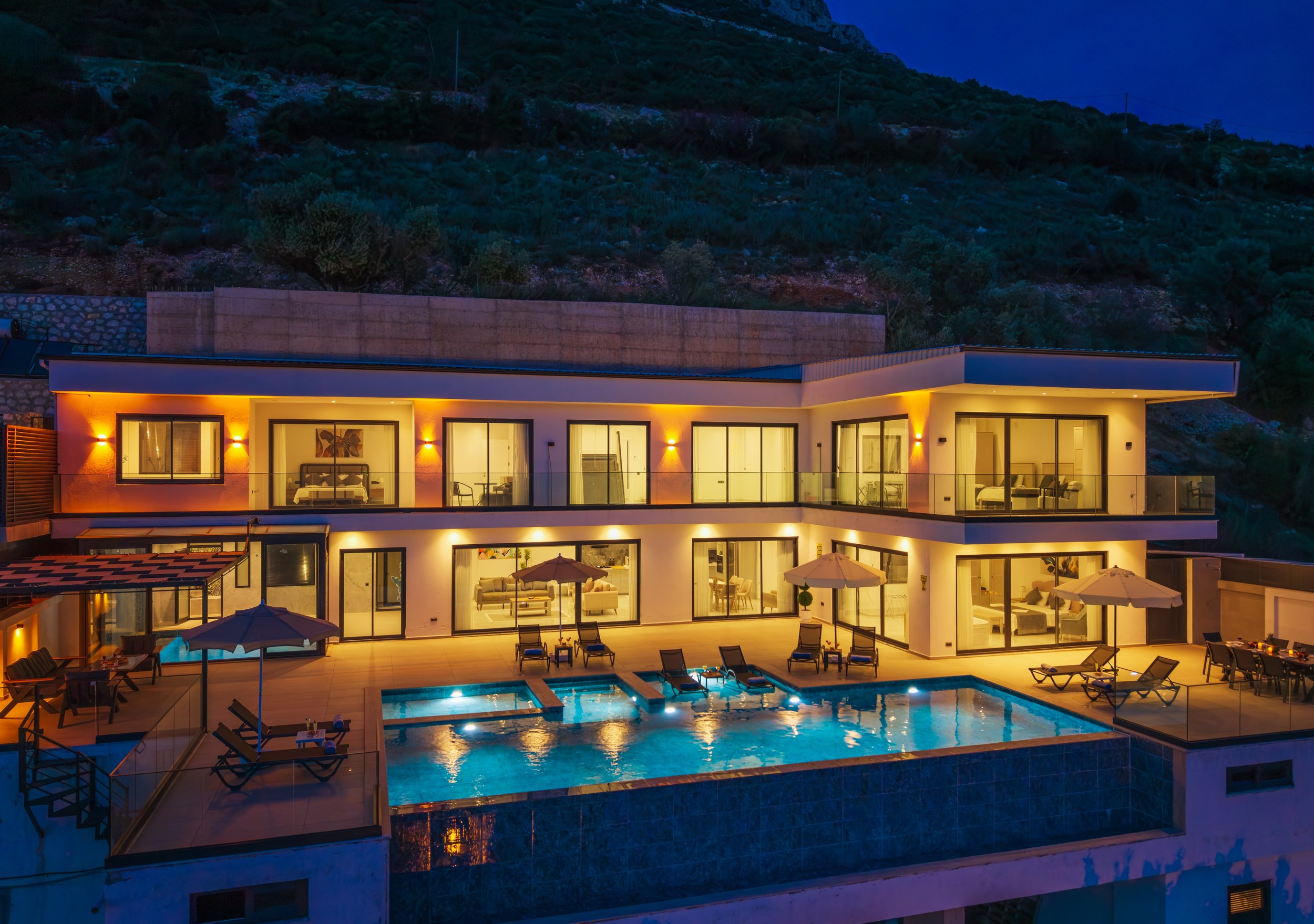 Night view of a modern multi-story house with illuminated interior and exterior, large glass windows, a swimming pool with lounge chairs, and a hillside covered with trees in the background.