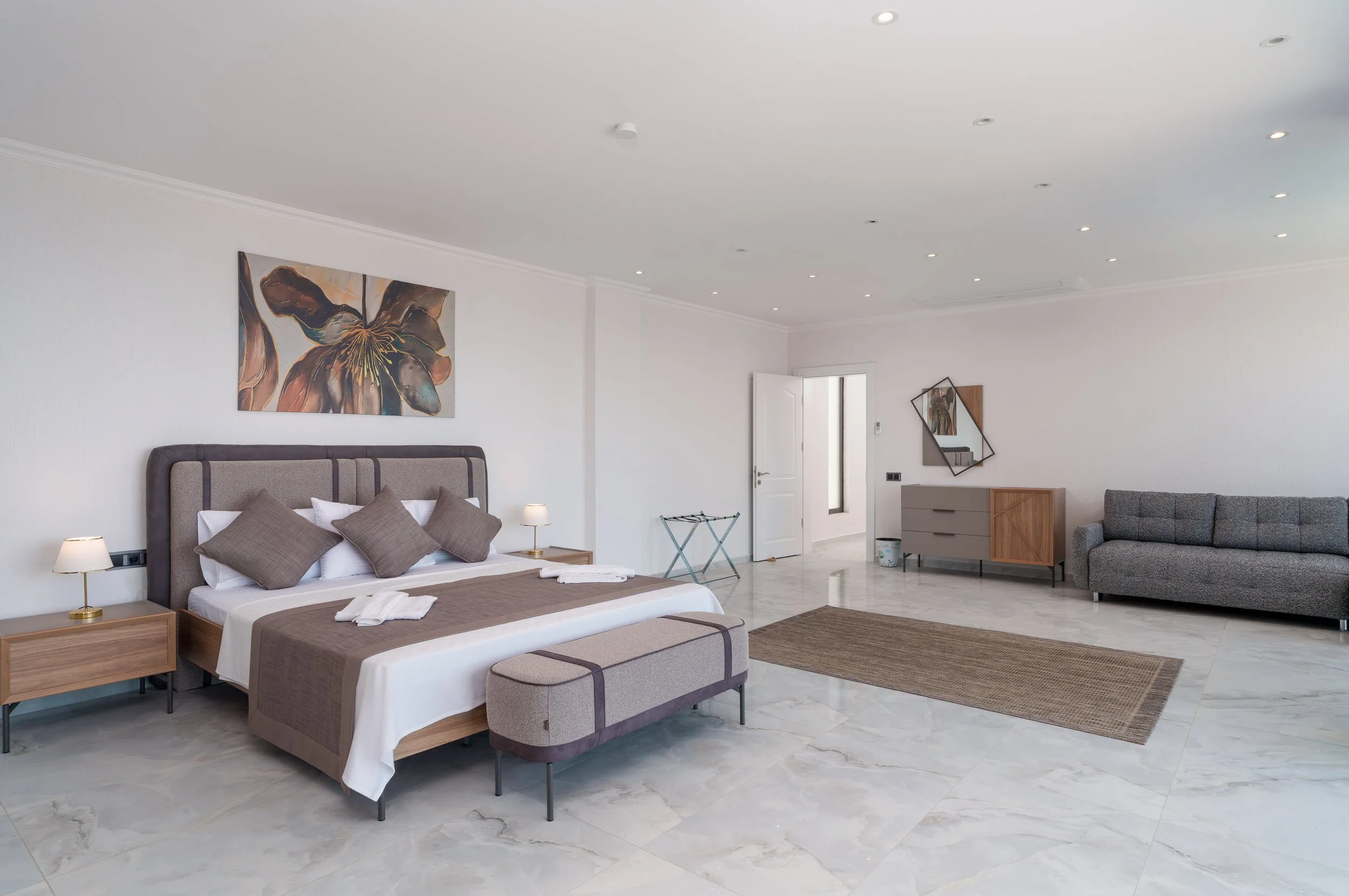 A modern bedroom with a large bed, beige headboard, gray and beige bedding, and multiple pillows. There are two nightstands with lamps on either side of the bed. On the wall above the bed, there is a large abstract flower painting. To the side, there