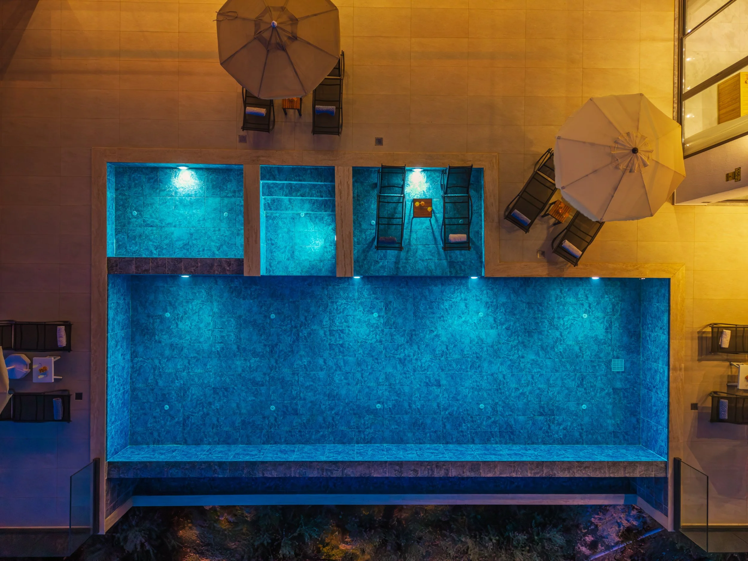 From an overhead view, a swimming pool area at night is illuminated with blue light, surrounded by beige tiles. Four black lounge chairs with rolled towels are arranged around the pool, two under beige umbrellas, with a small table between two chairs