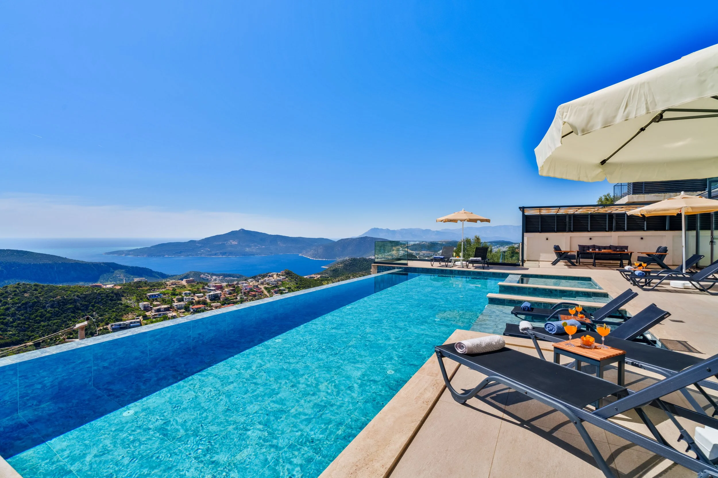 Luxury infinity pool overlooking a scenic landscape with mountains and ocean, outdoor lounge area with umbrellas and sunbeds, clear blue sky.