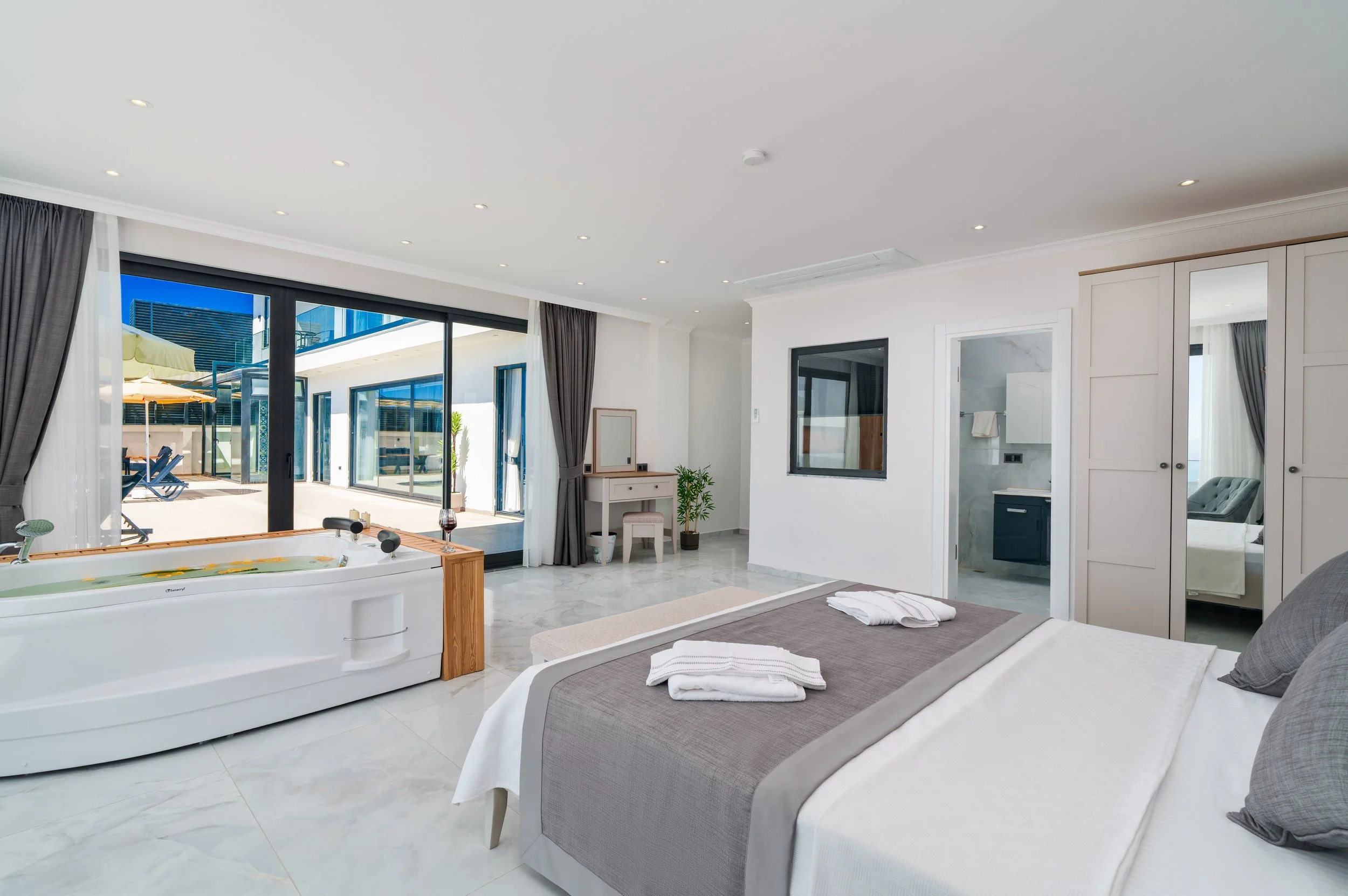 A modern bedroom with large sliding glass doors opening to an outdoor patio with umbrellas and lounge chairs, a Jacuzzi near the bed, white curtains, a small desk with a mirror, a potted plant, and a grey upholstered bed.