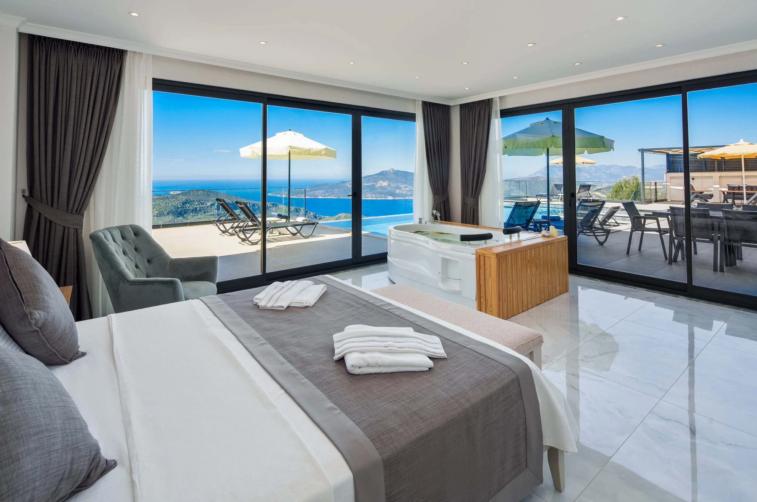 A bedroom with large sliding glass doors leading to a balcony with outdoor furniture and umbrellas, overlooking a scenic landscape with mountains and water.