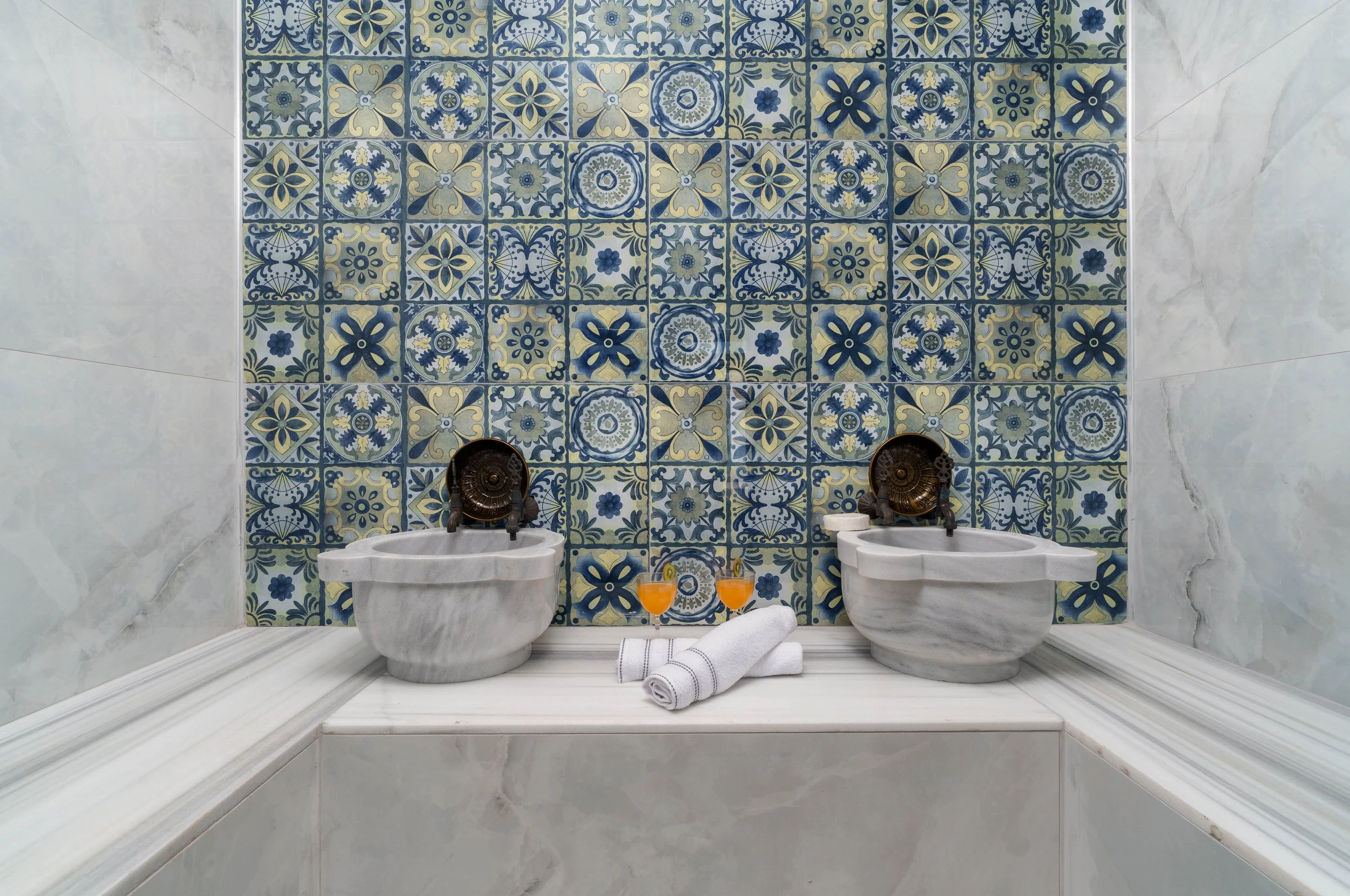 A luxurious bathroom with two white marble sinks, a vibrant blue and yellow patterned tiled wall, two glasses of orange juice, and neatly rolled white towels on the marble ledge.