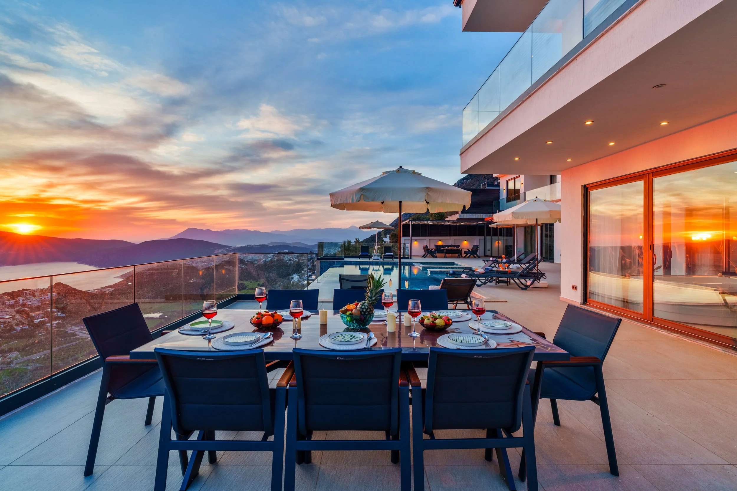 A dining table on a balcony at sunset with plates, glasses of red wine, and a fruit bowl, overlooking mountains and a cityscape.