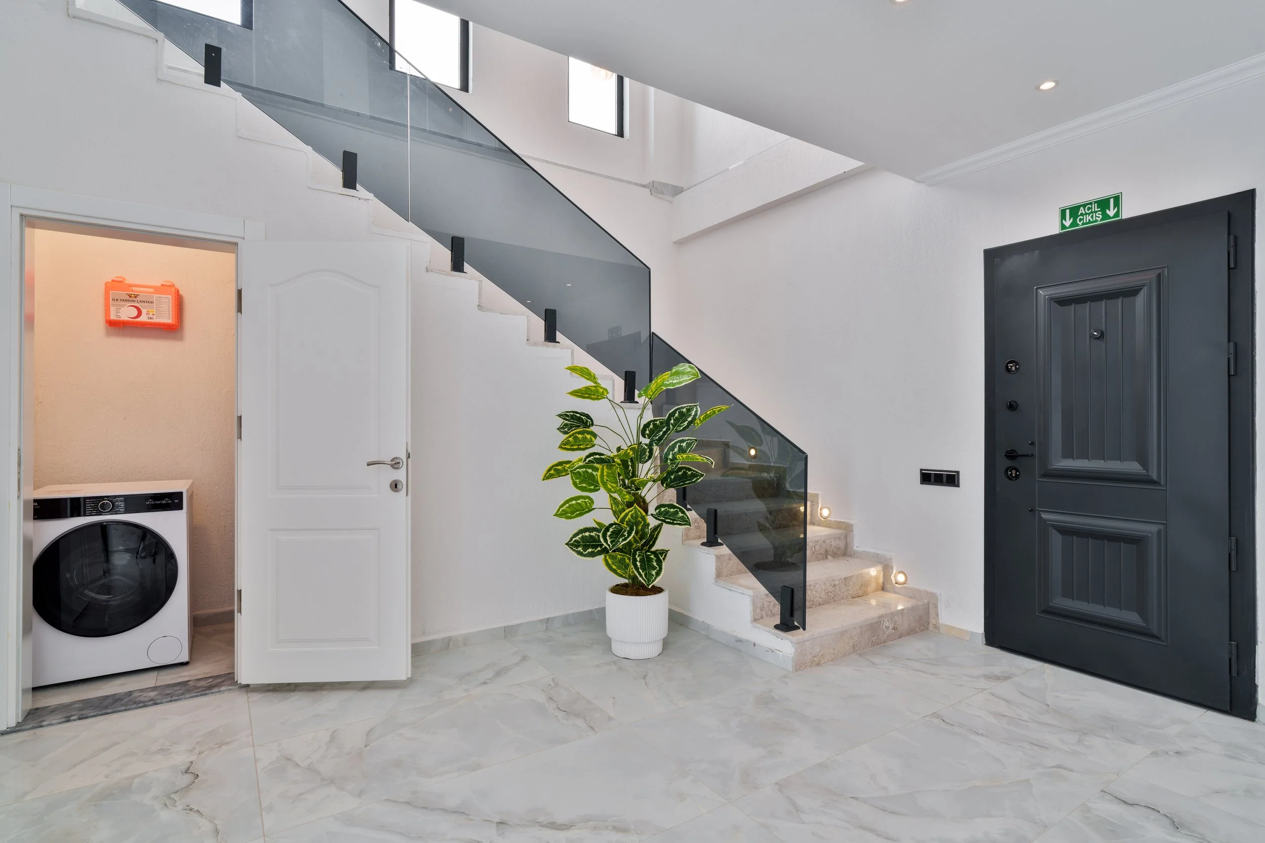 Interior of a modern house entryway with a staircase, potted plant, door, emergency kit, and laundry machine, featuring marble flooring and white walls.