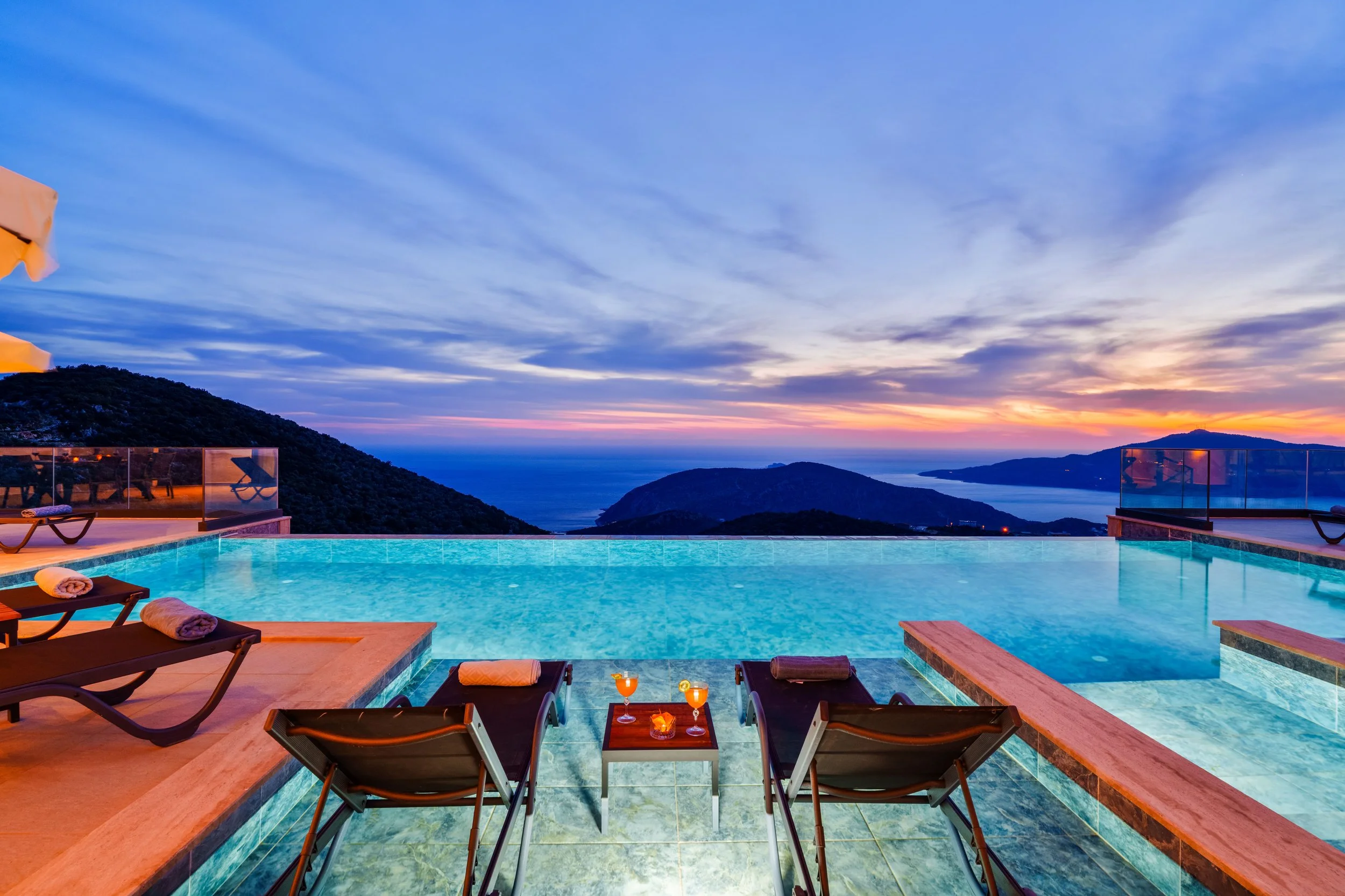 A scenic view of an infinity pool overlooking mountains and the ocean during sunset, with lounge chairs and drinks on a nearby table.