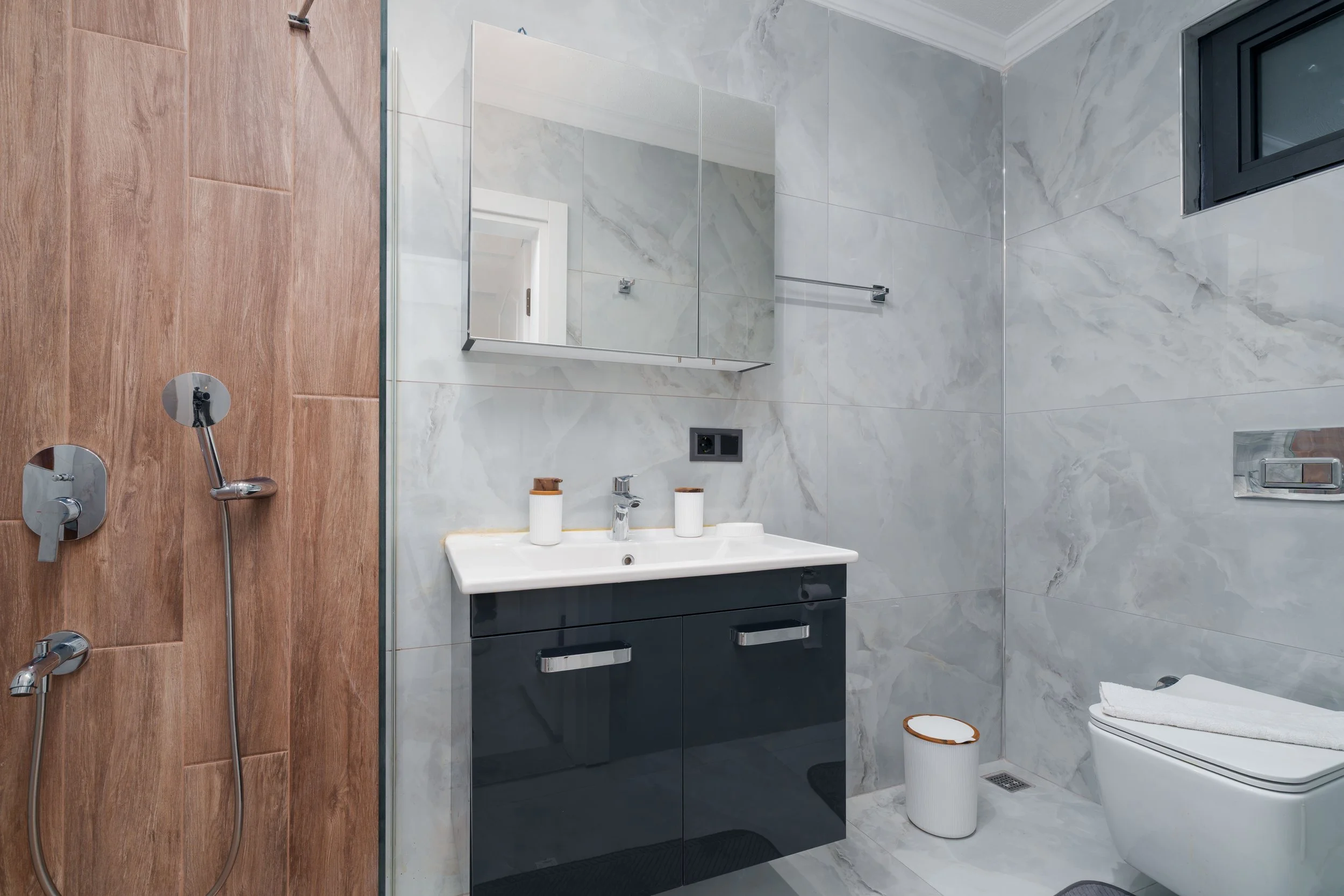 Modern bathroom with wooden and marble tile walls, a mirror, a sink vanity, a toilet, and a small window.