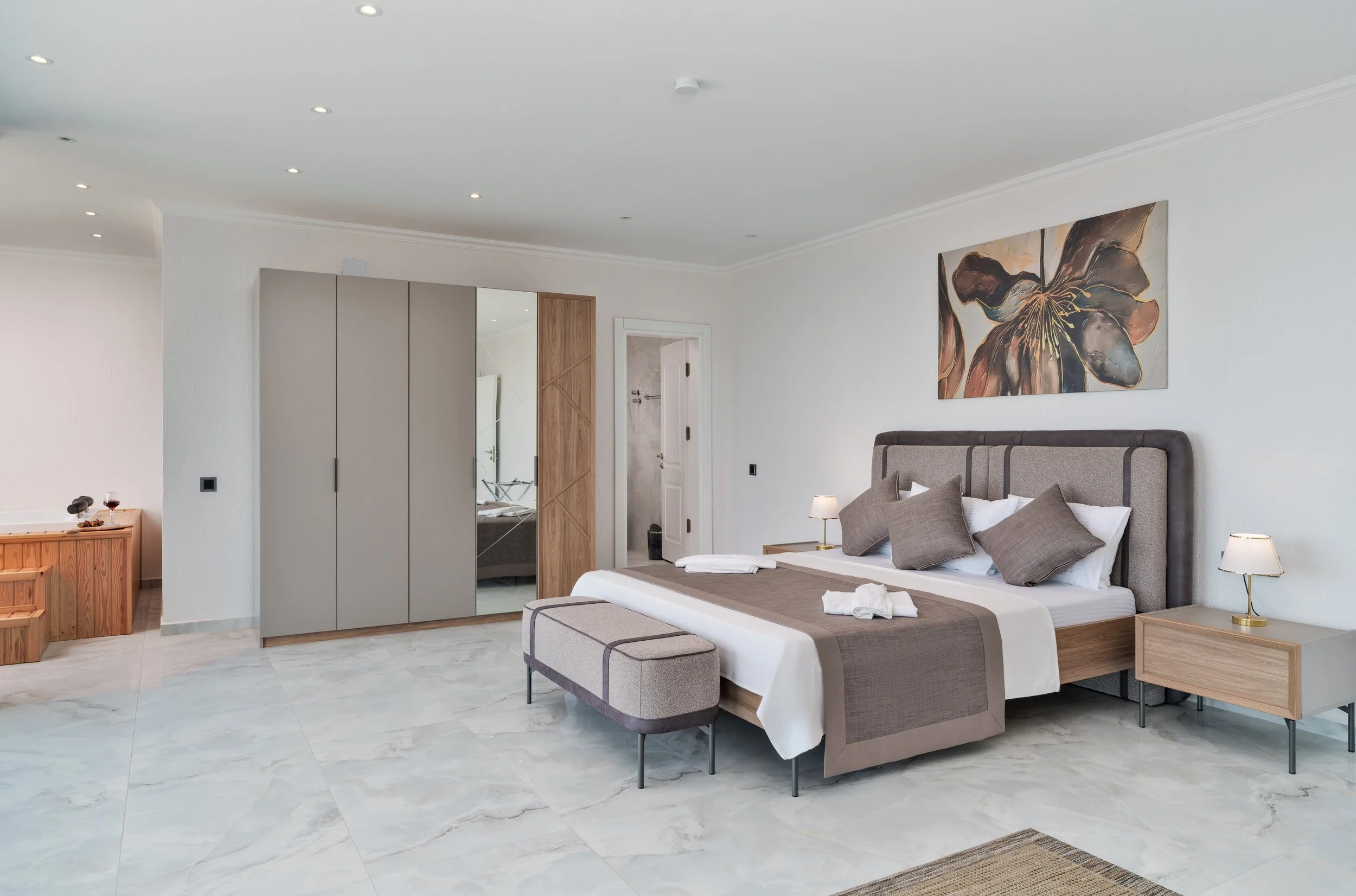 A modern bedroom with a gray upholstered bed, brown and gray pillows, a wooden side table with a lamp, abstract floral artwork on the wall, a wardrobe with mirrored doors, and a small bench at the foot of the bed. The room has white walls, a white ce