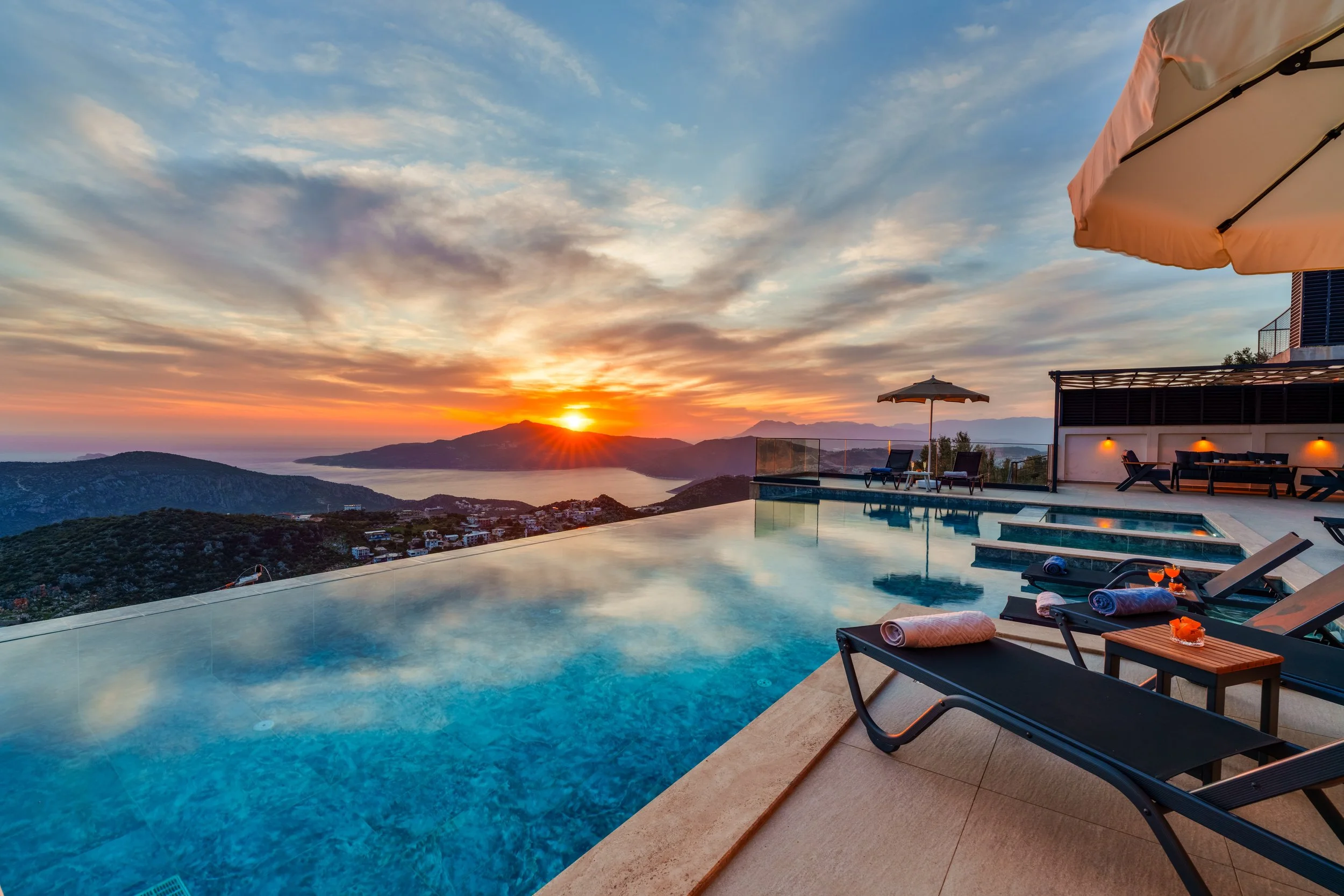 A luxurious infinity pool overlooking mountains and a body of water at sunset. there are lounge chairs, towels, and drinks on the deck, with a large umbrella and a modern house edge.