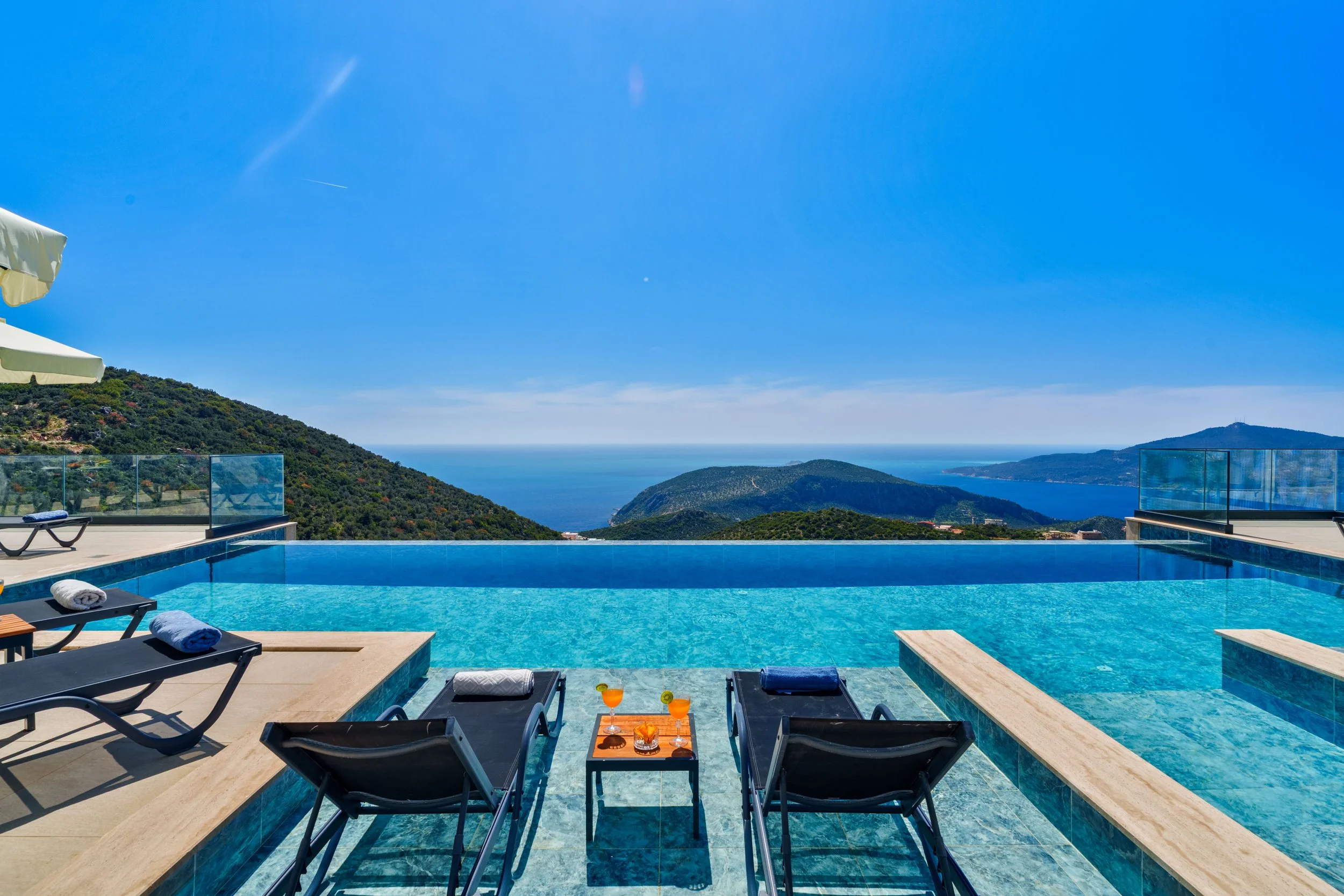 View of a swimming pool with lounge chairs and towels, overlooking green hills, ocean, and blue sky.