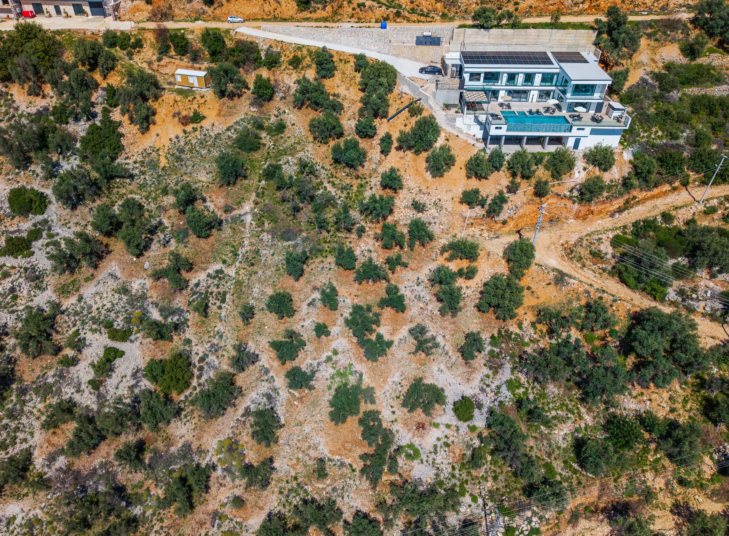Aerial view of a modern white house with a swimming pool on a hillside with trees and dry terrain.