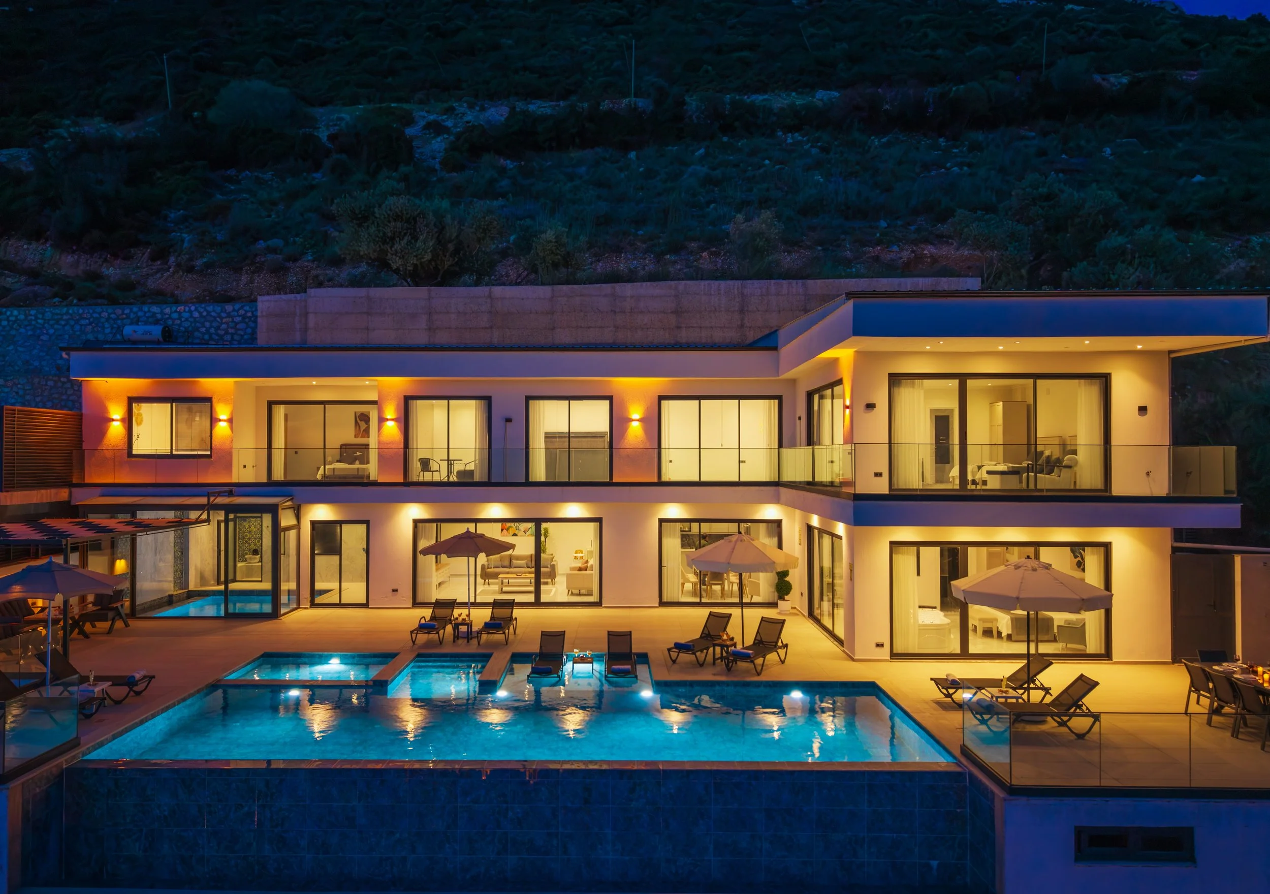 Modern multi-level house with large glass windows illuminated at night, featuring an outdoor swimming pool and patio area with lounge chairs and umbrellas, set against a hillside with vegetation.