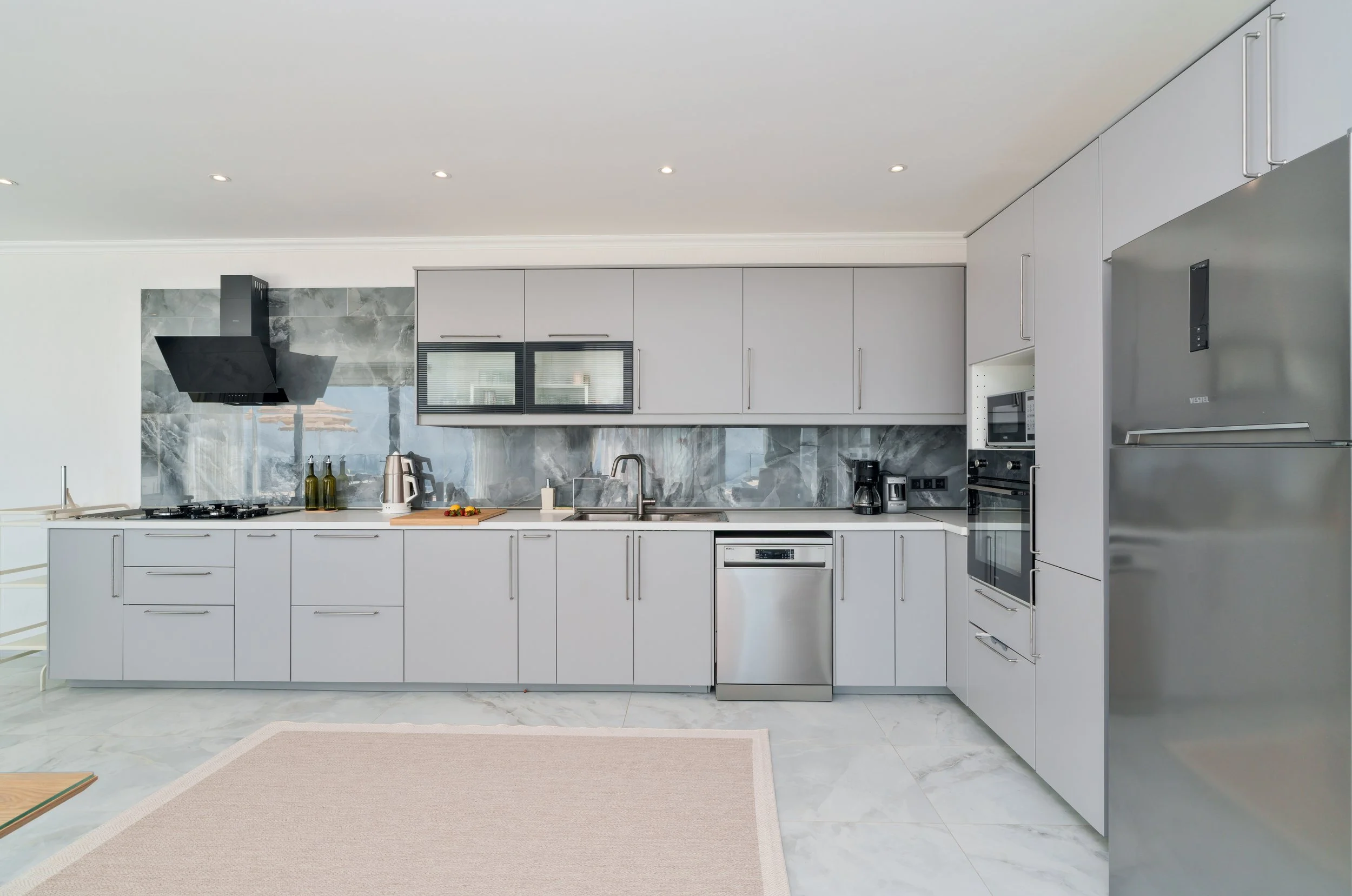Modern kitchen with white cabinetry, stainless steel appliances, marble backsplash, black range hood, and a beige rug on a tiled floor.