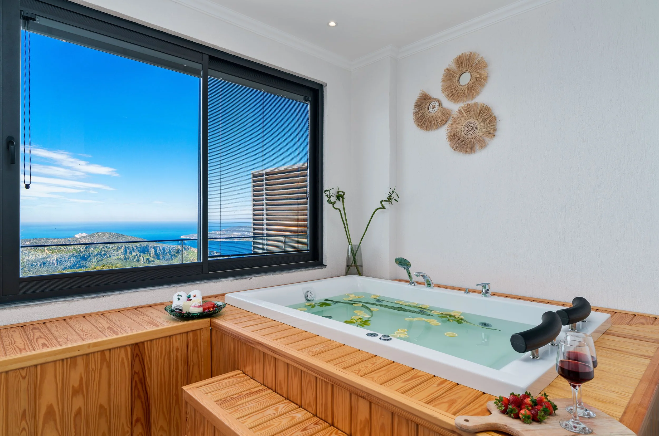 A bathroom with a large window offering a view of the ocean and hills. There is a white jacuzzi tub filled with water and floating lemon slices. On the edge of the tub, there is a glass of red wine, strawberries, and a wooden paddle with strawberries