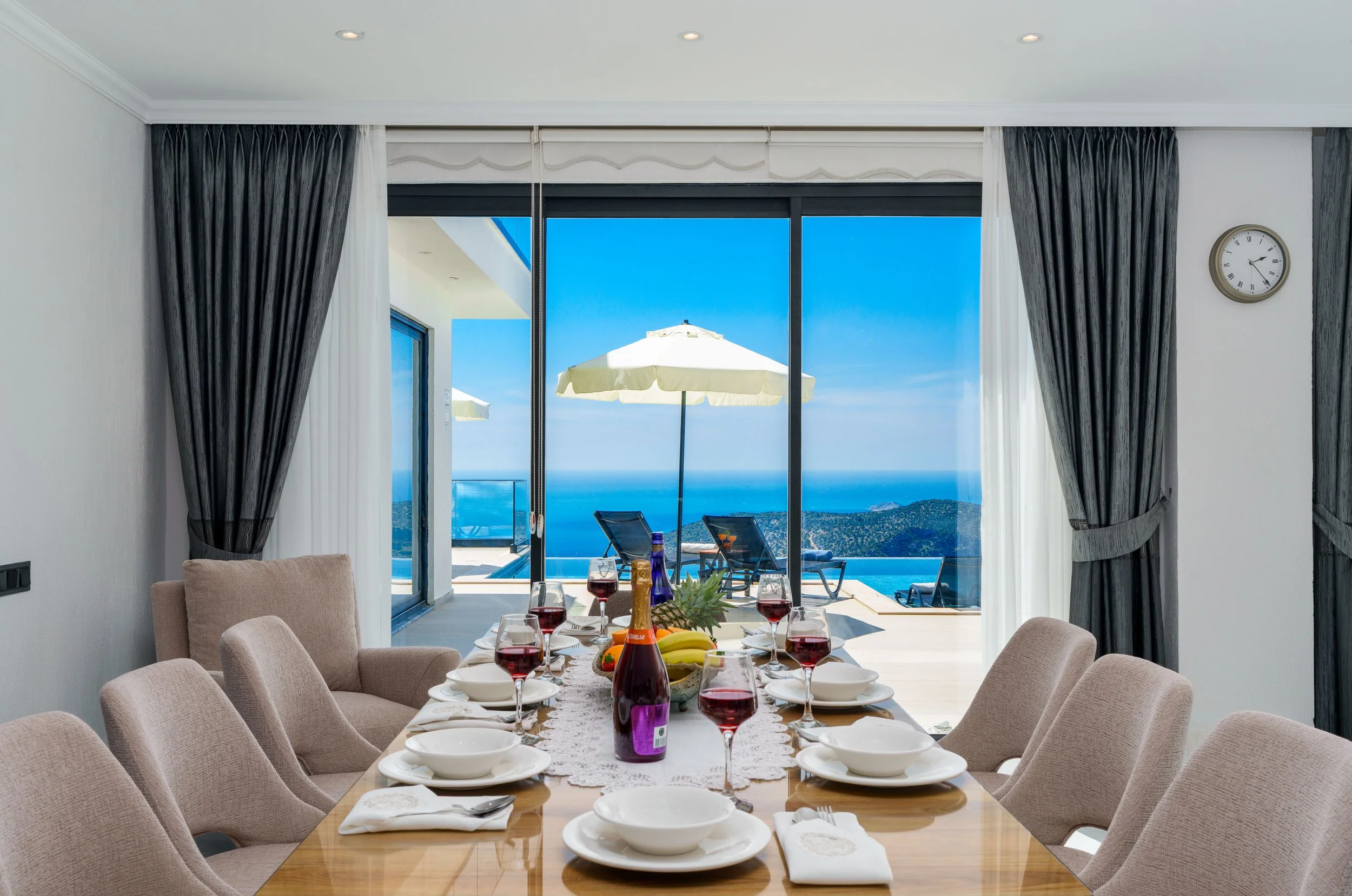 A dining room with a large window revealing a view of the ocean and blue sky. The table is set with white plates, wine glasses filled with red wine, and a centerpiece of fruit and bottles. There are beige chairs around the table, and the room has dar