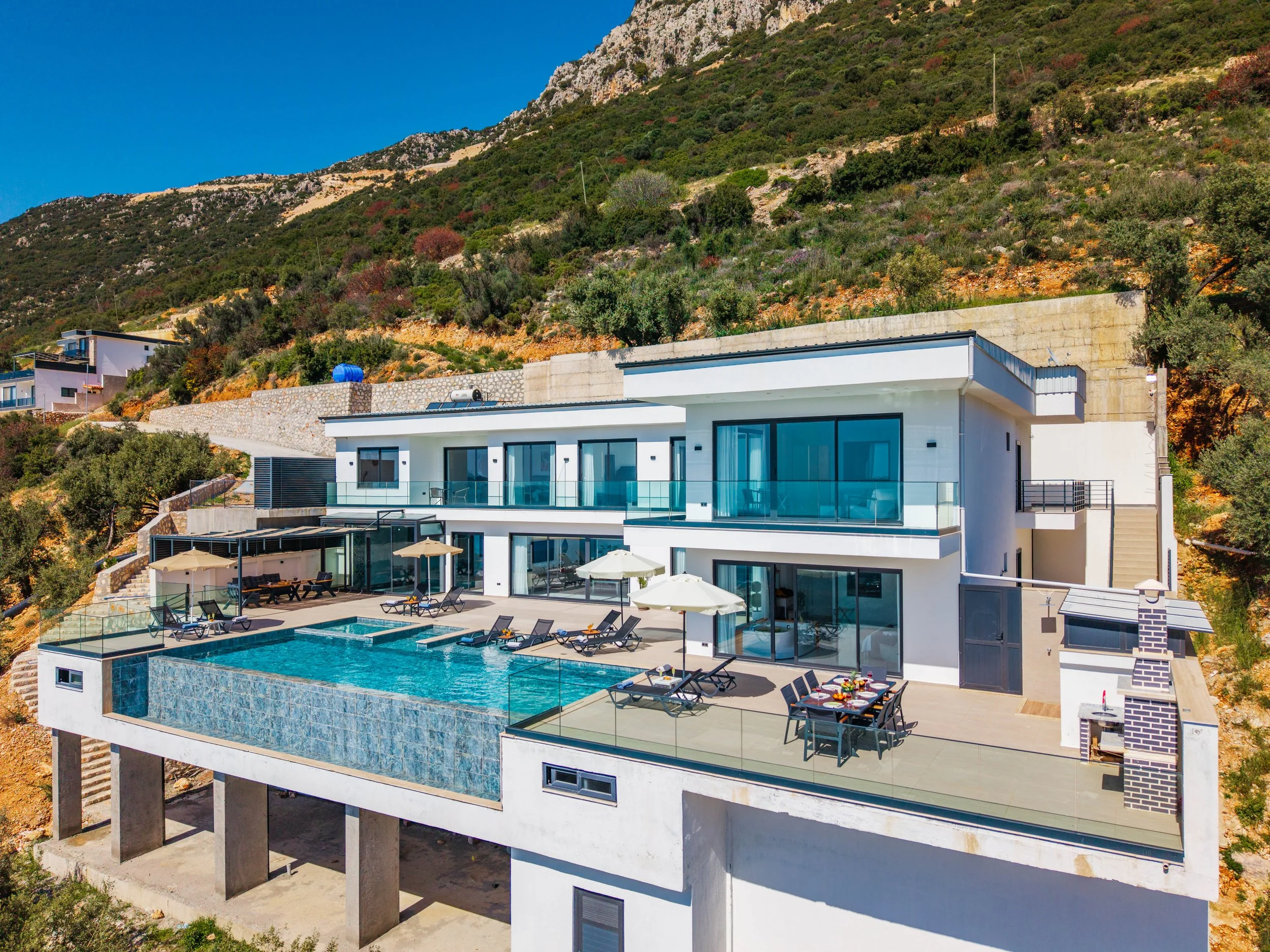 Modern white multi-story house with large glass windows and outdoor swimming pool, situated on a hillside with lush green vegetation and rocky terrain in the background.