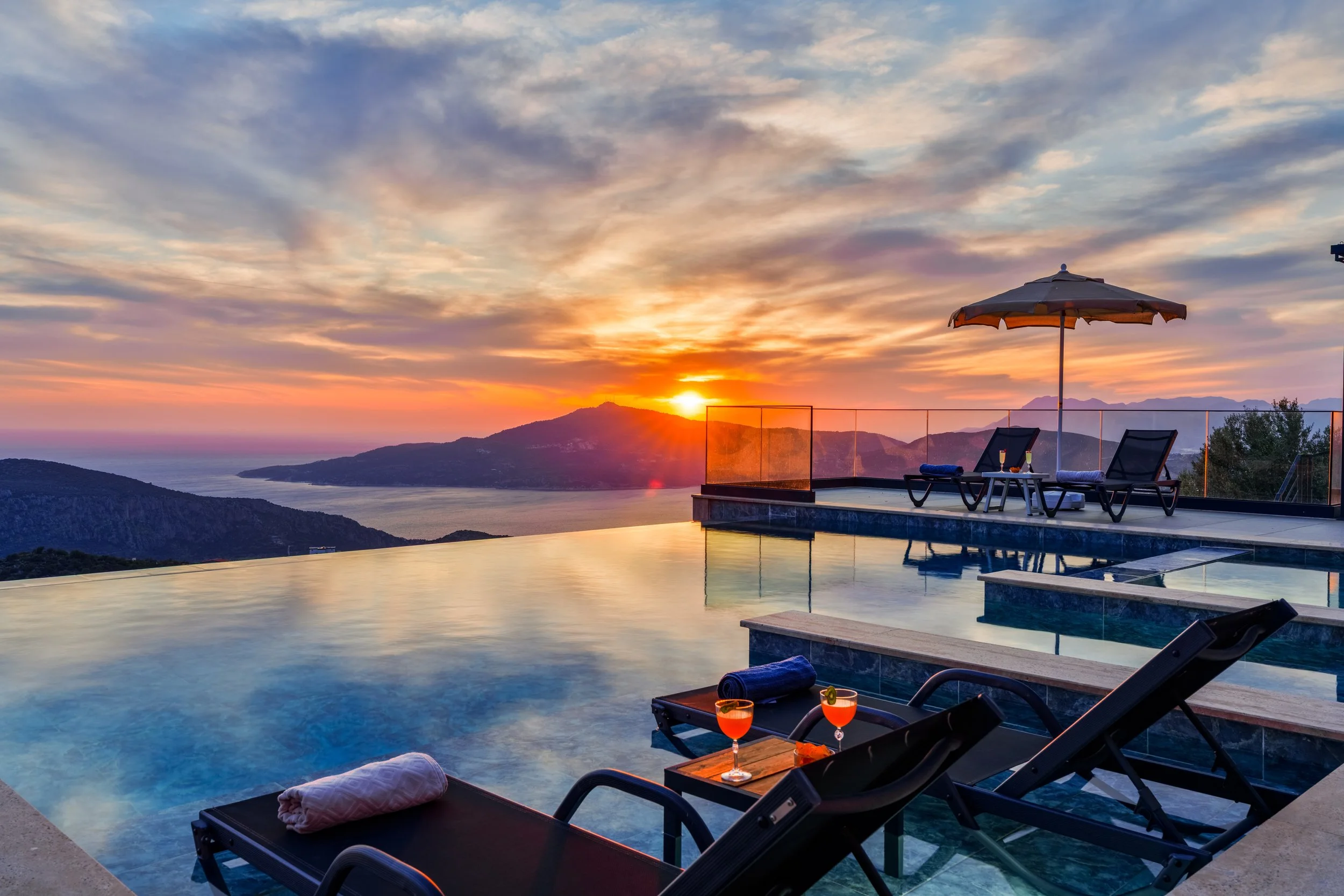 A scenic sunset view over mountains and a body of water, with a luxurious infinity pool in the foreground. Poolside lounge chairs and tables with drinks are visible, along with an umbrella and towels.