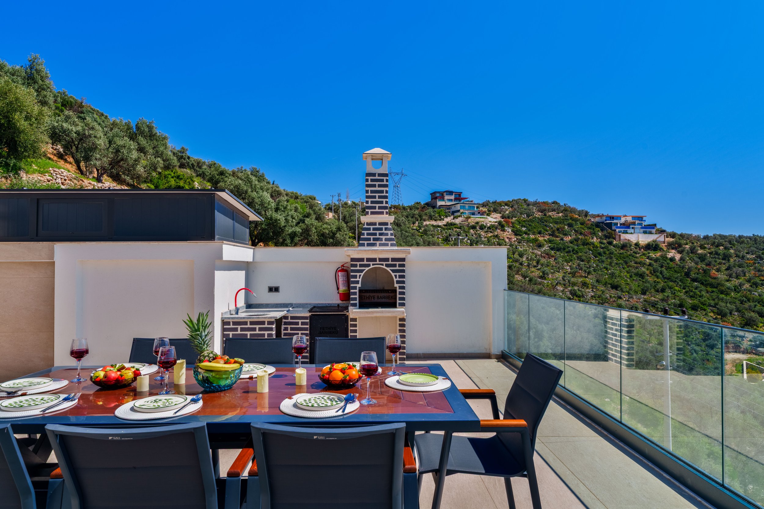 An outdoor dining table set with plates, wine glasses, and bowls of fruit on a terrace with a glass railing and a view of hillside houses against a blue sky.