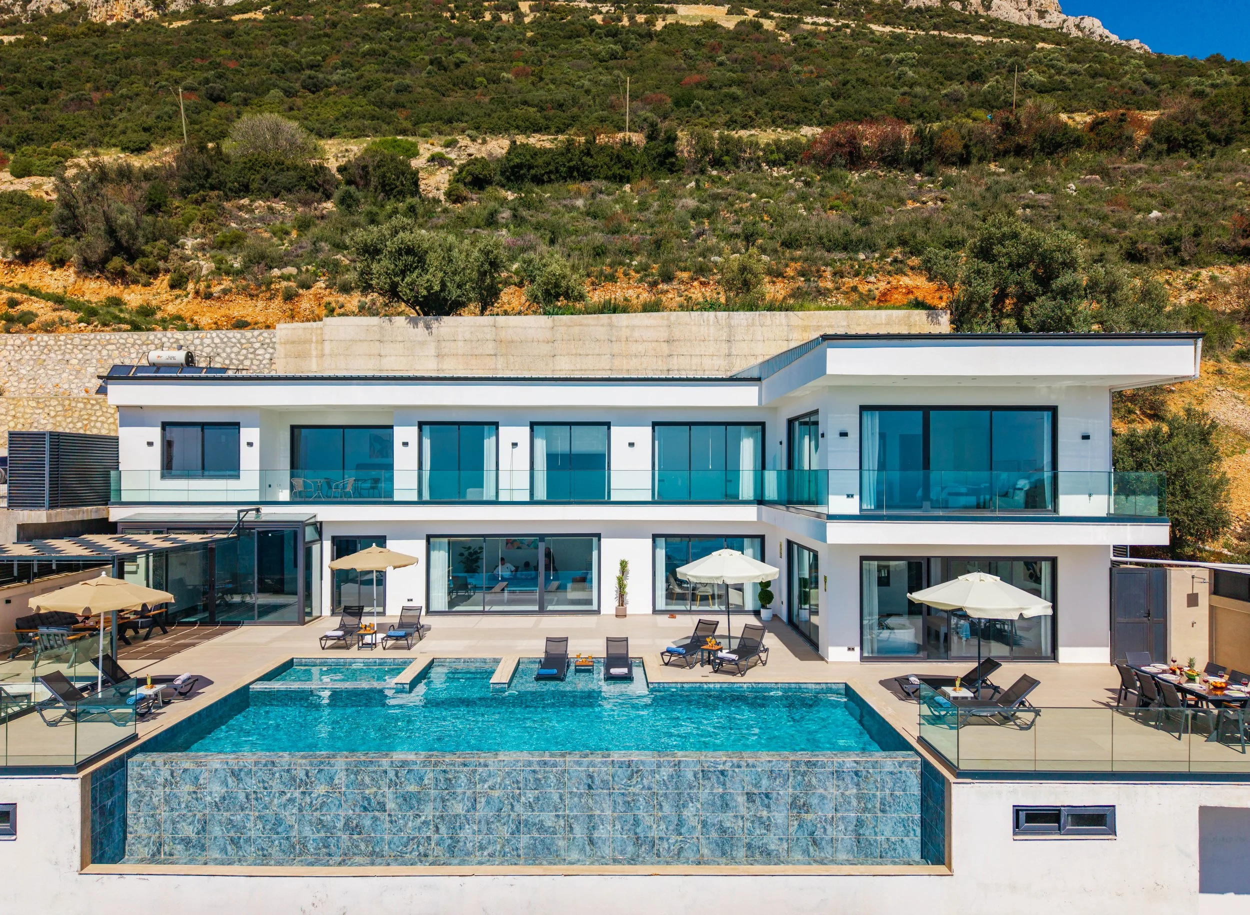 Modern white house with large glass windows, outdoor pool area with lounge chairs and umbrellas, set against a hillside with green shrubs and trees.