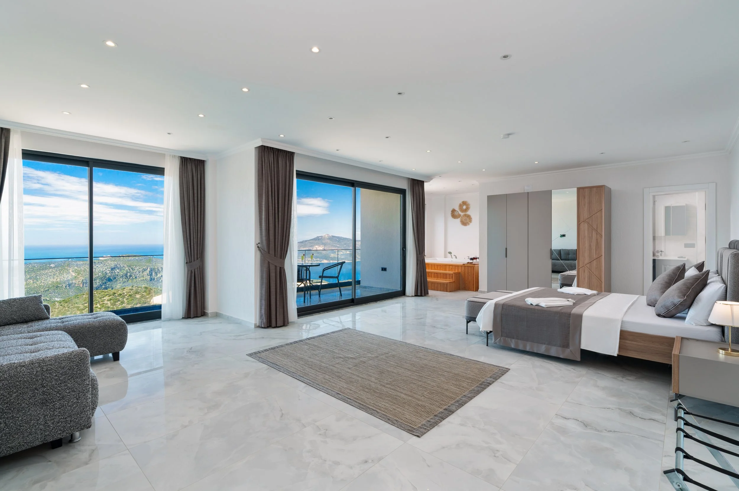 Modern living room with marble floors, large windows showing a scenic view of hills and the ocean, a bed with white and gray bedding, a gray sofa, a beige rug, and a balcony with outdoor furniture.