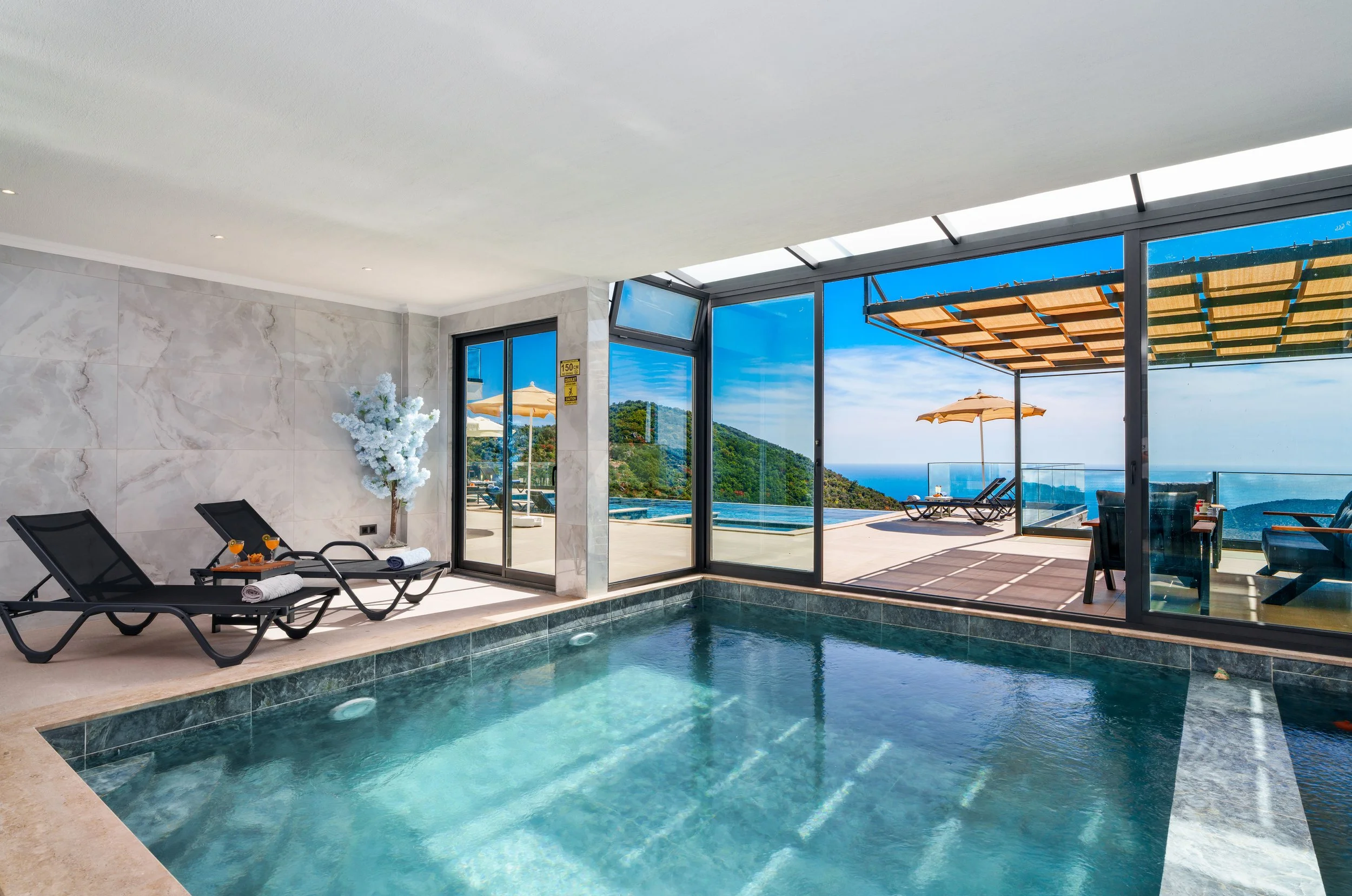 Indoor pool area with a view of an outdoor patio, umbrellas, deck chairs, and scenic hills in the background through large glass windows.