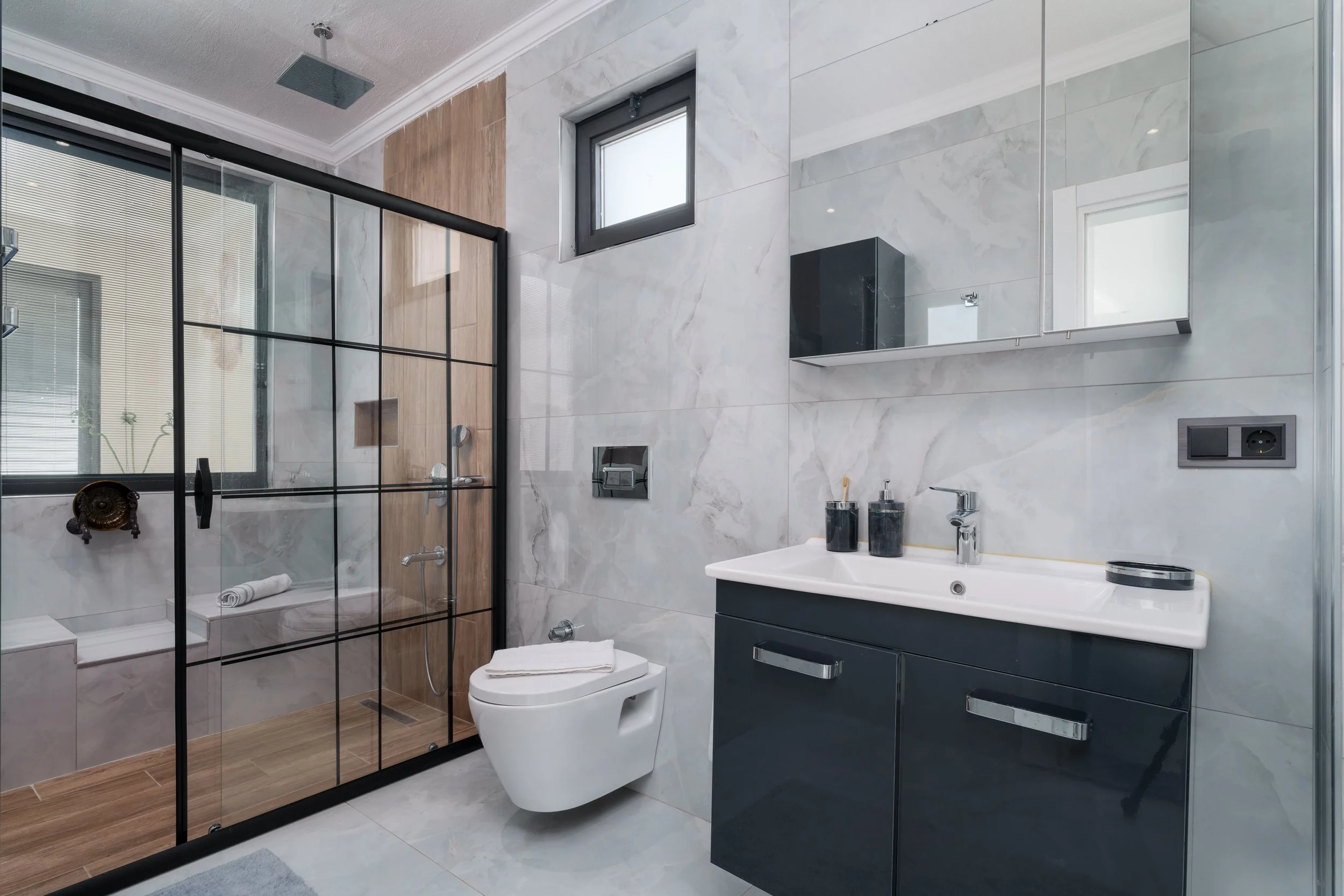 Modern bathroom with a glass-enclosed shower, a white toilet, a dark gray vanity with a mirror, a small window, and gray marble tile walls.
