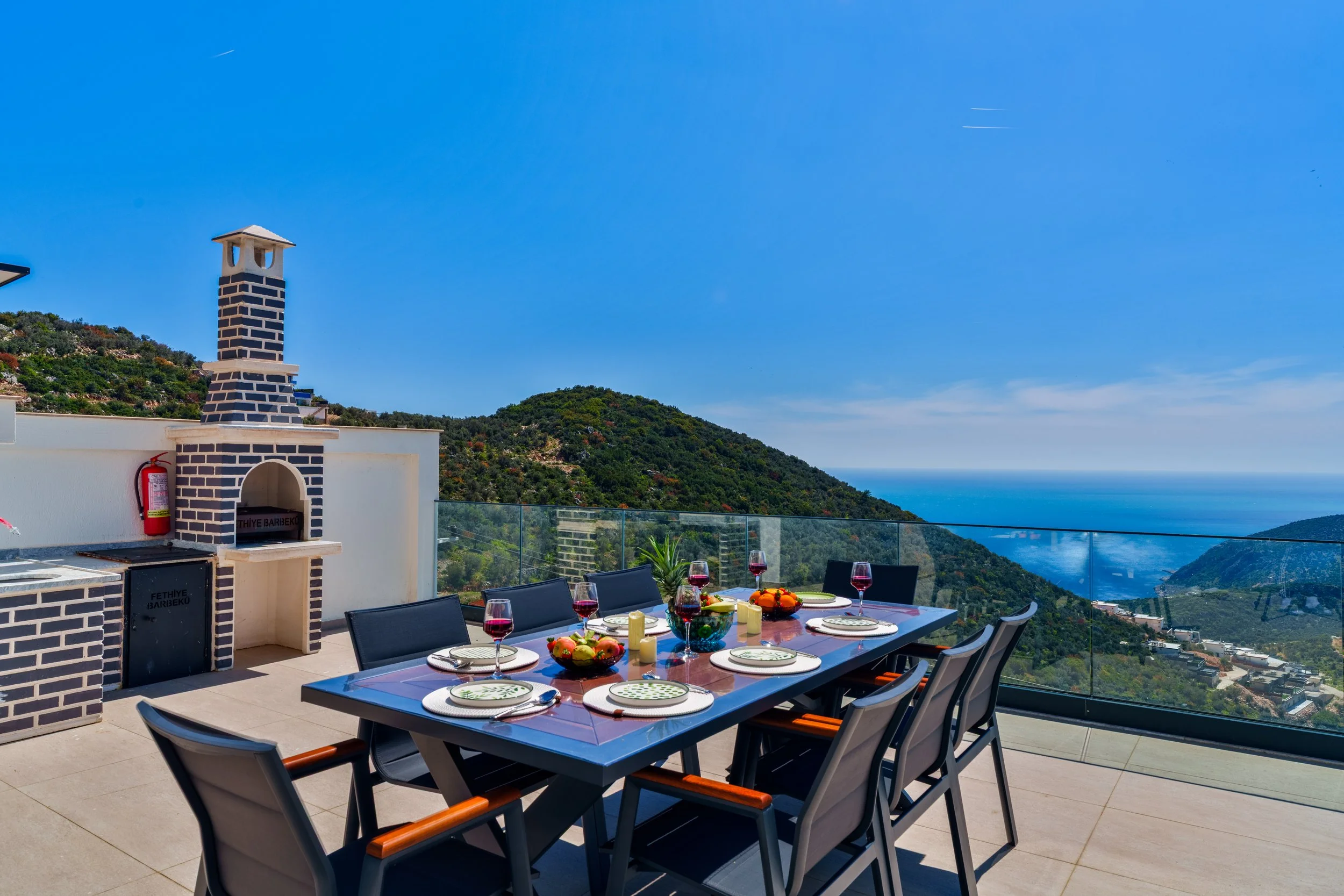 Outdoor dining setup on a terrace with a view of green hills and the ocean, featuring a table with plates, glasses of red wine, and a decorative centerpiece, with a brick barbecue and a fire extinguisher in the corner.